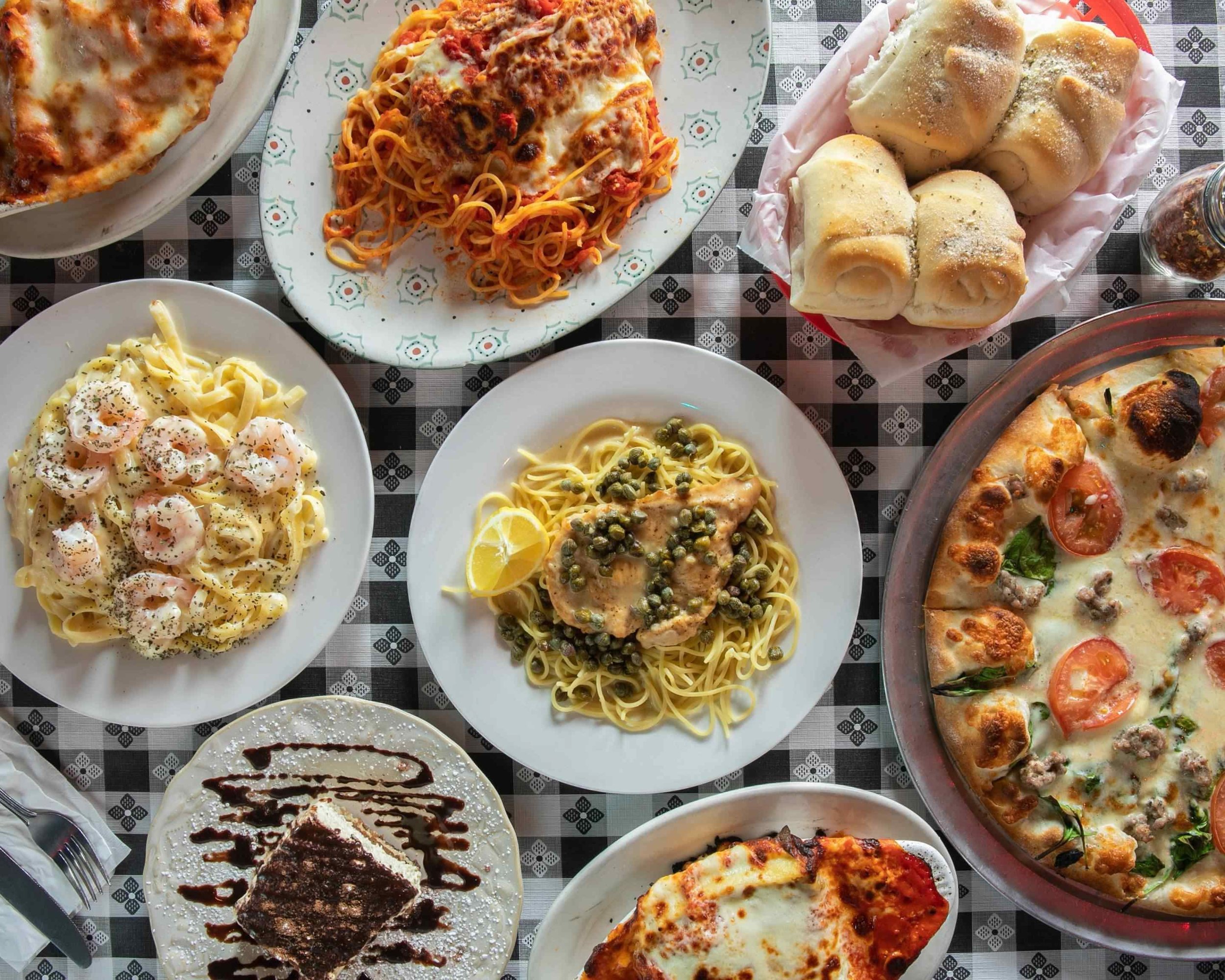 A variety of Italian dishes on a checkered tablecloth, including salads, pasta, pizza, bread rolls, and a dessert with chocolate drizzle.