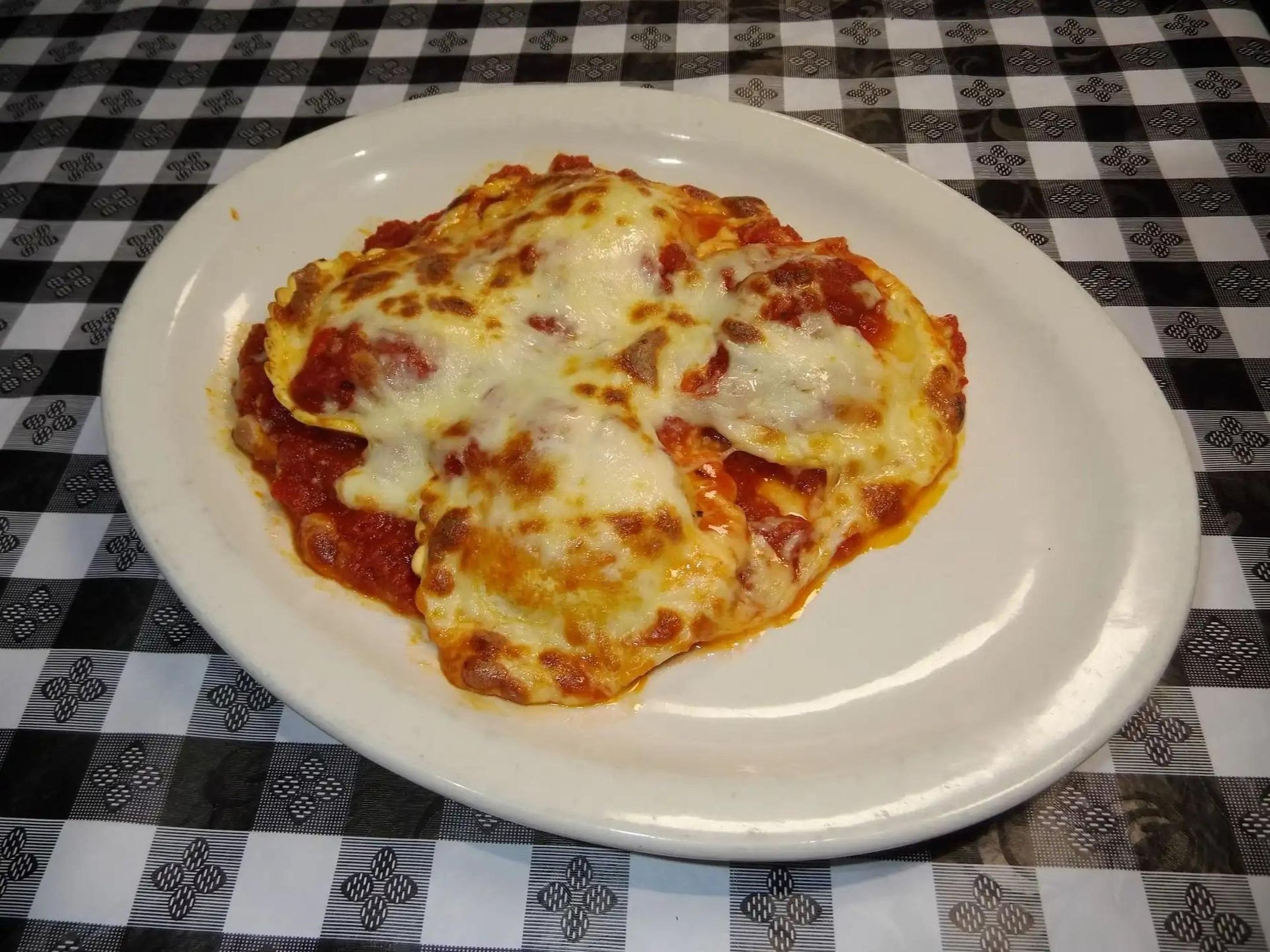 Slice of pizza with melted cheese, tomato sauce, and toppings on a white plate, placed on a black and white checkered tablecloth.