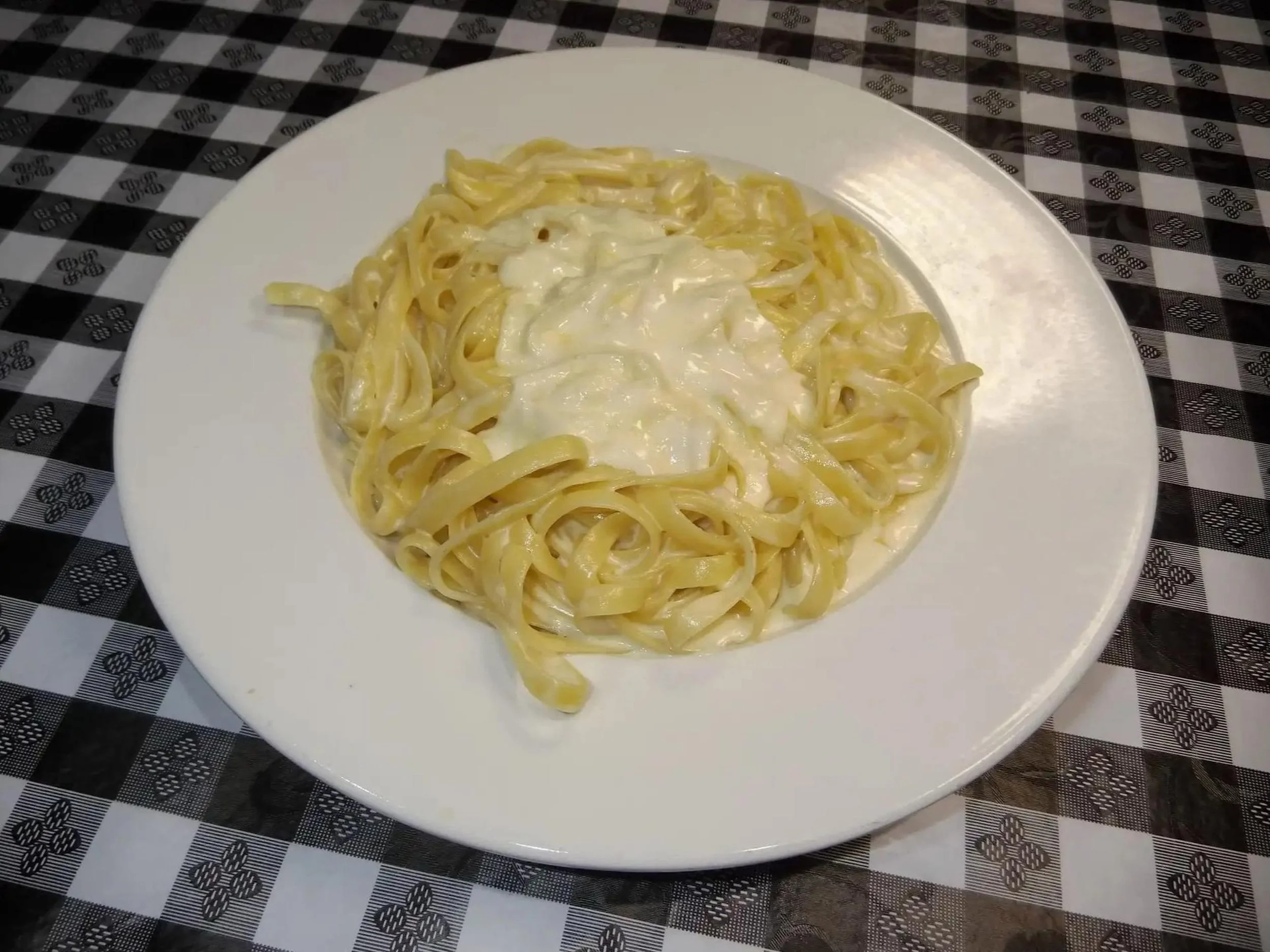 A white plate with cooked fettuccine pasta in a creamy white sauce, placed on a checkered black and white tablecloth.