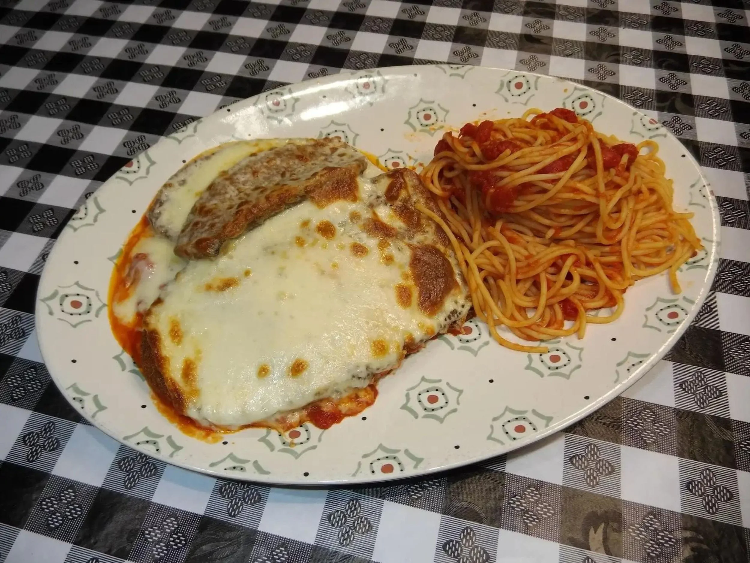 A plate of baked pasta with melted cheese, accompanied by a serving of spaghetti with tomato sauce, on a decorative plate placed on a black and white checkered tablecloth.