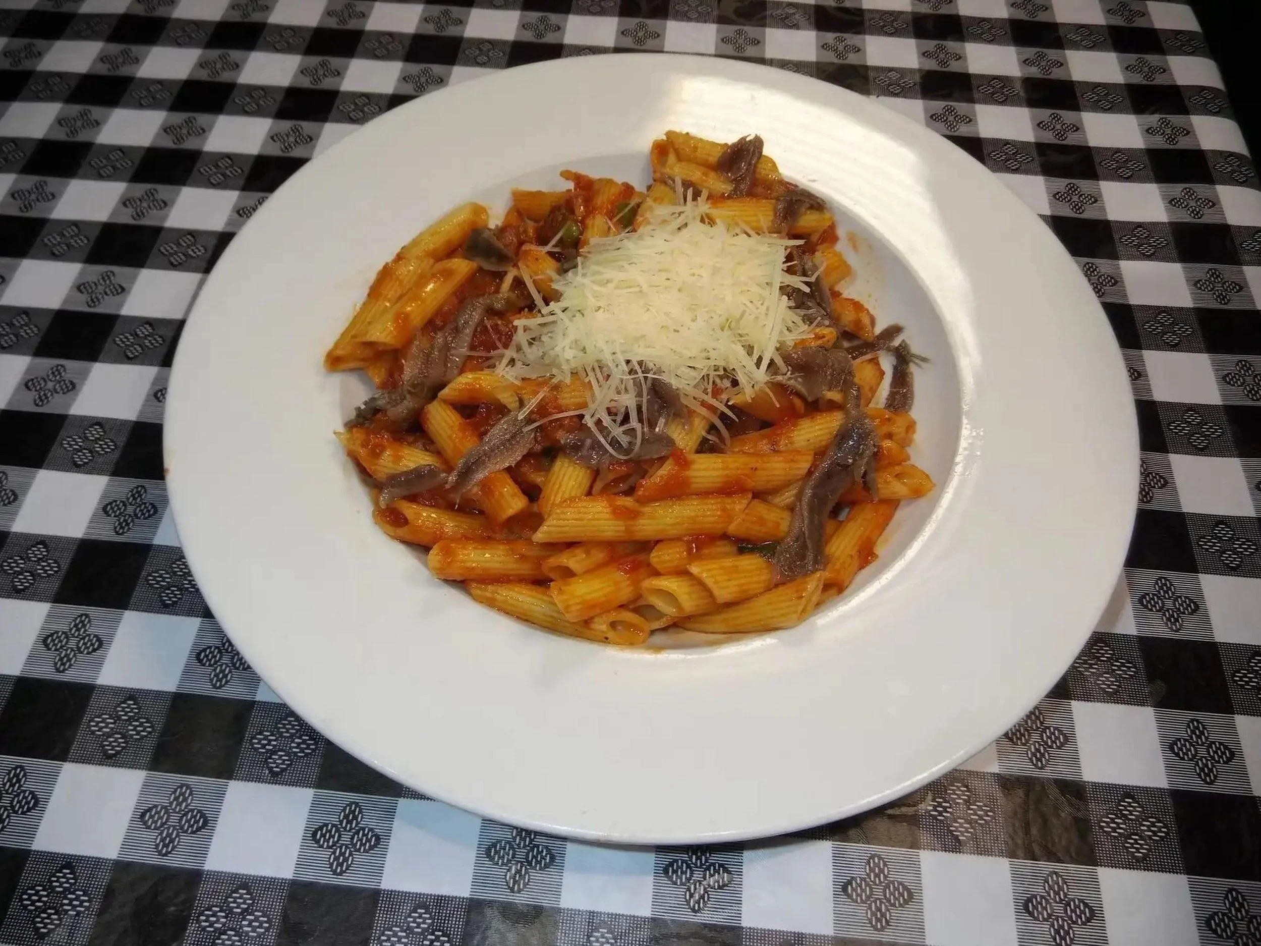 A white plate of pasta with tomato sauce, topped with shredded cheese, and garnished with small pieces of meat, placed on a black-and-white checkered tablecloth.