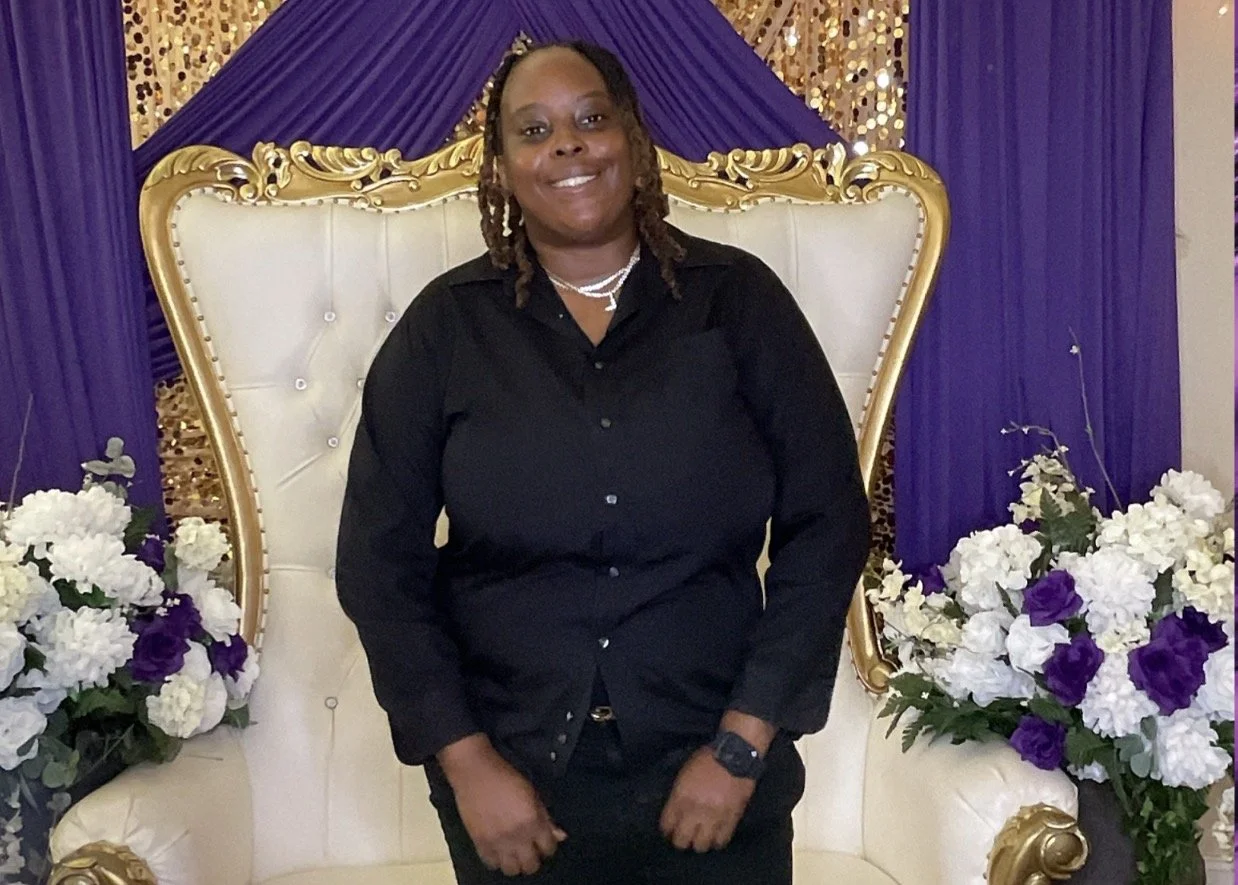 A smiling woman with dreadlocks wearing a black shirt and jewelry, standing on a white and gold ornate throne with floral arrangements of white and purple flowers on each side, in front of a purple and gold backdrop.