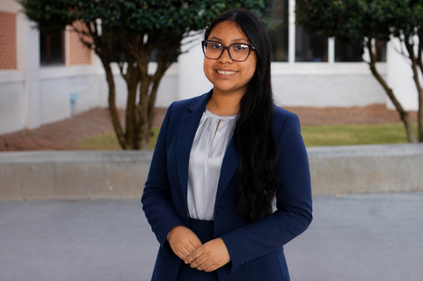 Young woman with long black hair and glasses, wearing a navy blue blazer and white blouse, standing outdoors in front of trees and a building, smiling.