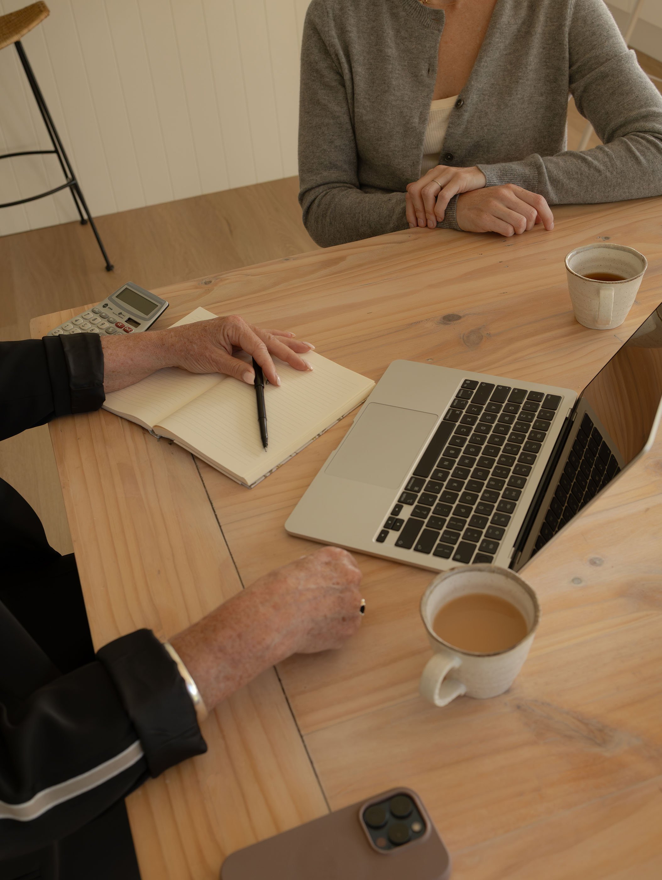 A wooden table with a silver laptop, a smartphone, a notebook, a remote control, a cup of coffee, and two people sitting at the table talking. One person has a hand resting on the notebook, and the other person's hands are clasped on the table.