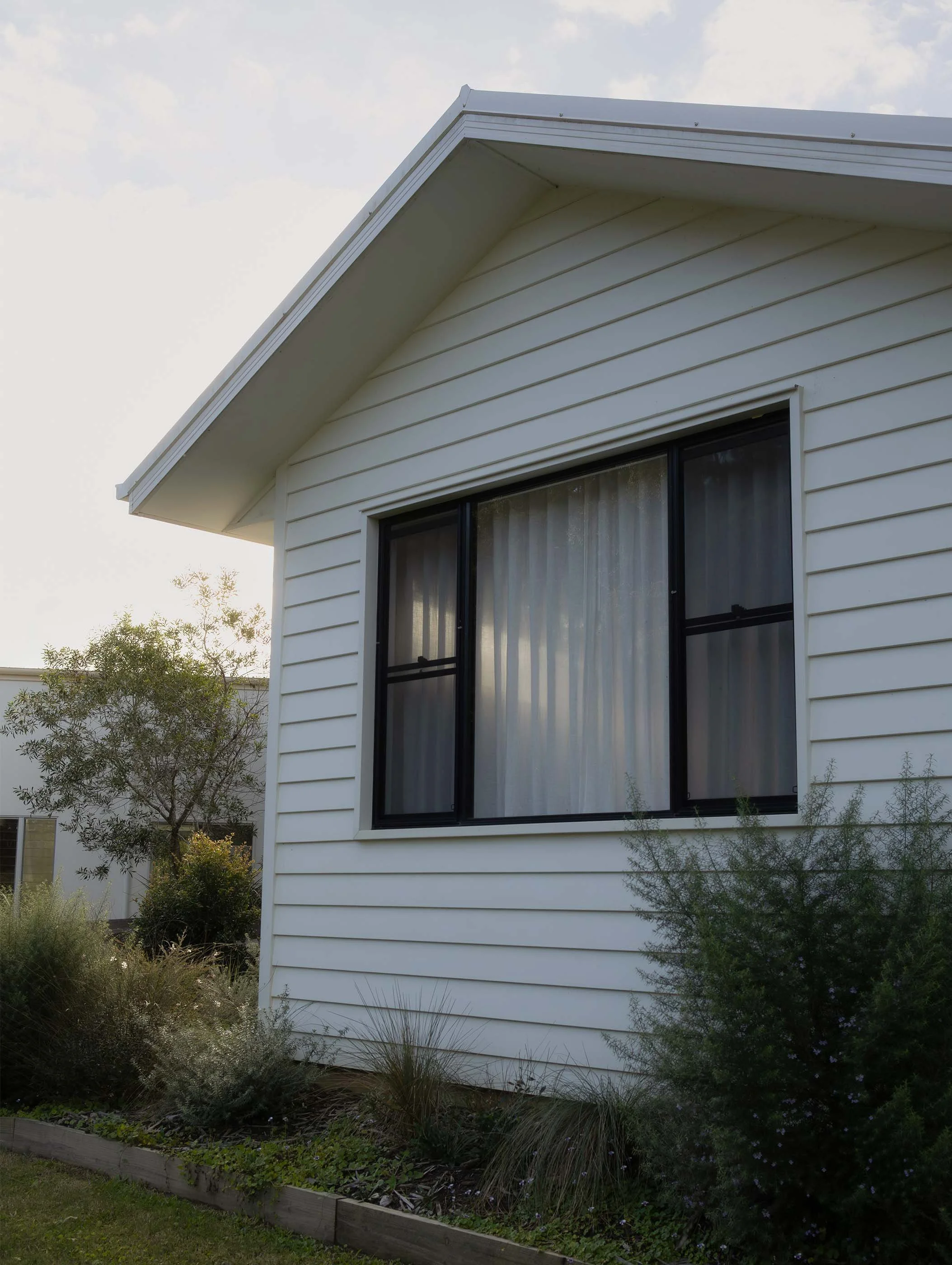 Corner of a modern white house with horizontal siding and large sliding glass door, surrounded by landscaped garden with bushes and flowering plants, under a partly cloudy sky.