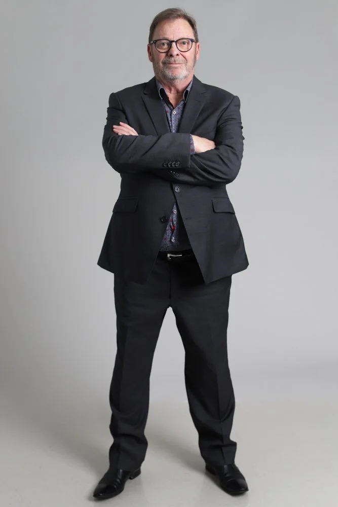 A man with glasses, a beard, and wearing a dark suit stands with arms crossed against a neutral gray background.