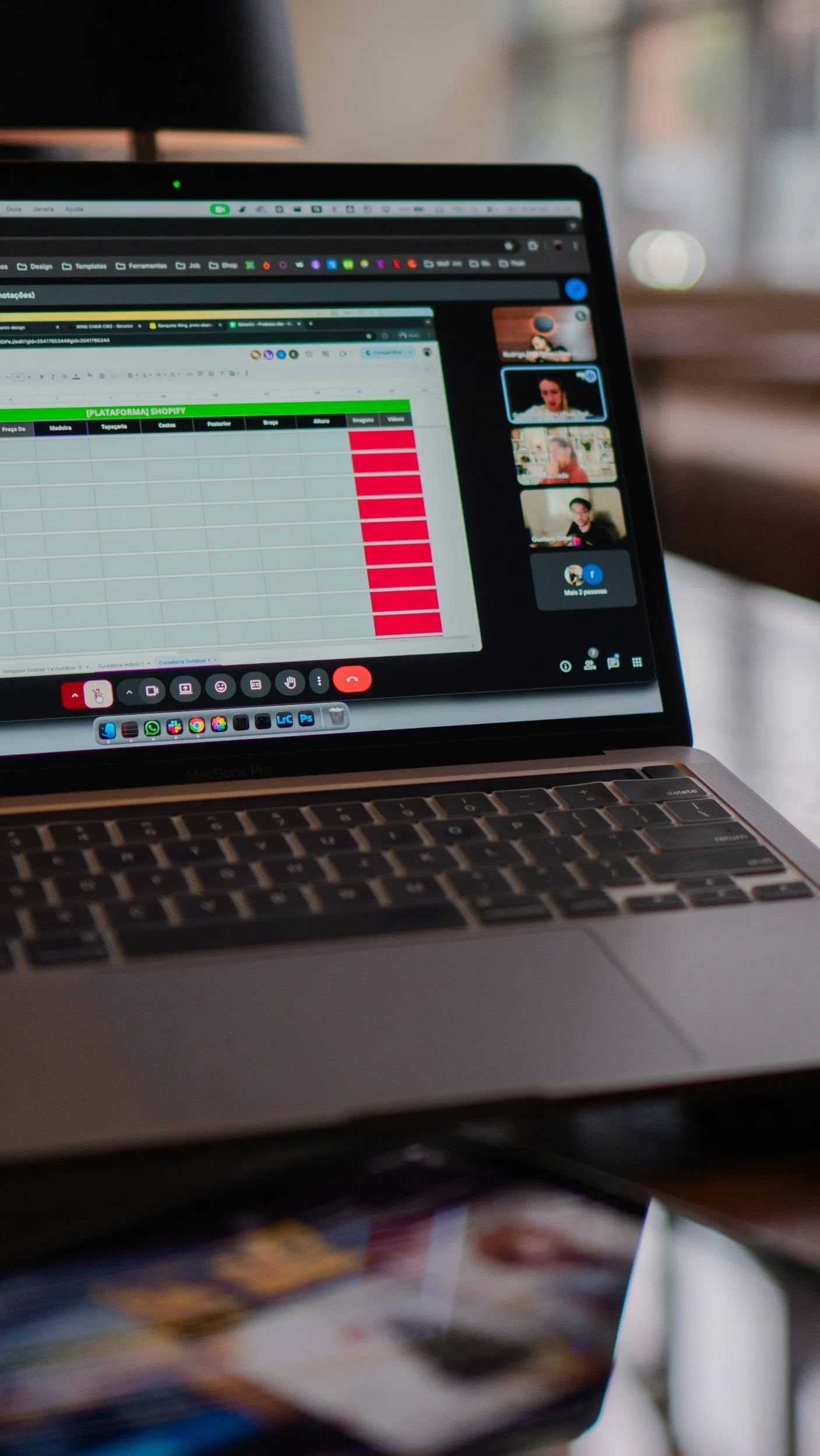 A laptop screen showing a spreadsheet and a video call with four participants. The laptop is on a desk near a window with natural light.