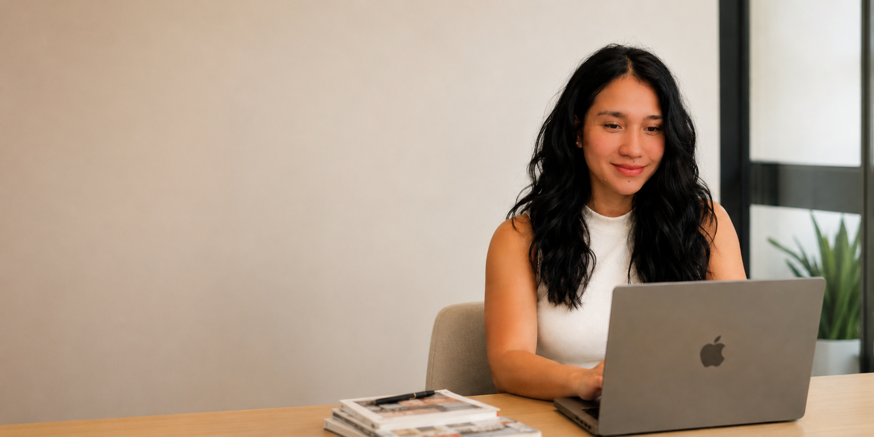 A woman with long black wavy hair sitting at a wooden table with a silver Apple MacBook laptop in front of her, looking at the screen and smiling. There's a stack of magazines and a pen on the table. In the background, a large window and a green plant are visible.