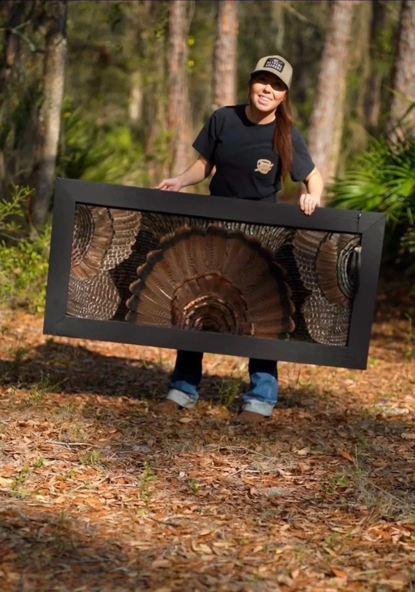 A woman standing outdoors in a wooded area, holding a framed piece of art that depicts a dinosaur fossil.