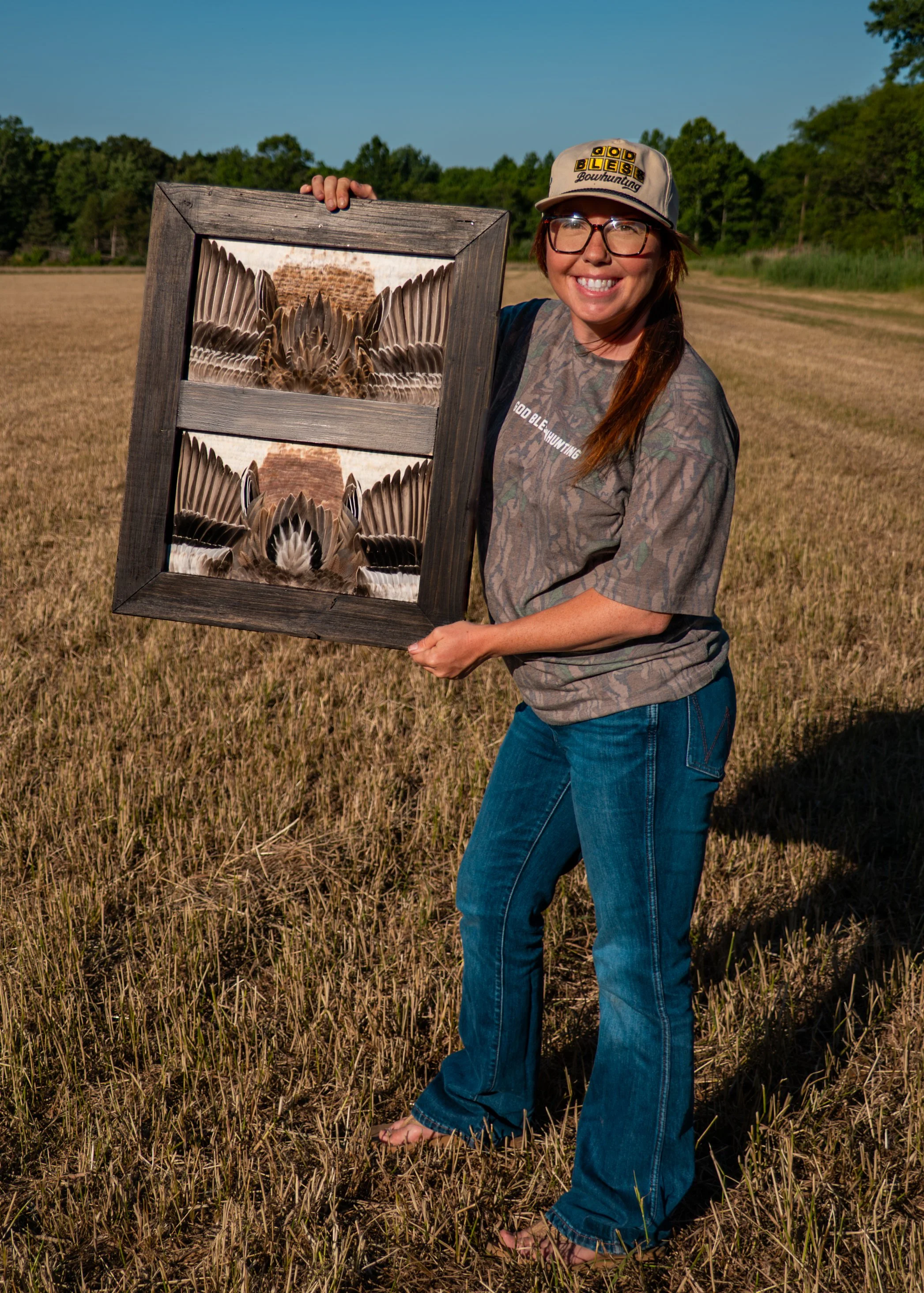 Woman smiling outdoors holding a framed photo of a turkey with feathers spread out in a field.