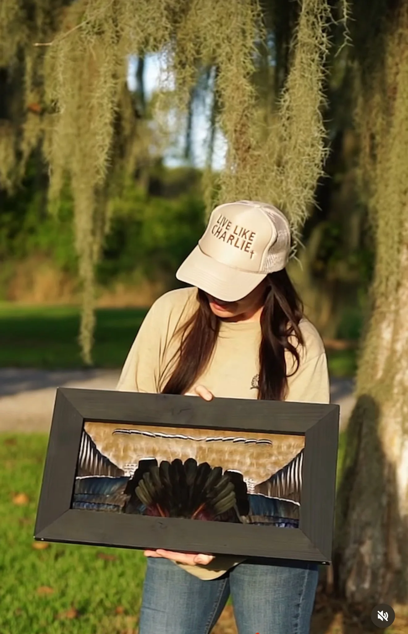 A woman wearing a beige baseball cap with the words 'Live Like Charlie' stands outdoors holding a framed artwork with a black circular design. She is wearing a beige t-shirt and jeans, with trees and hanging moss in the background.