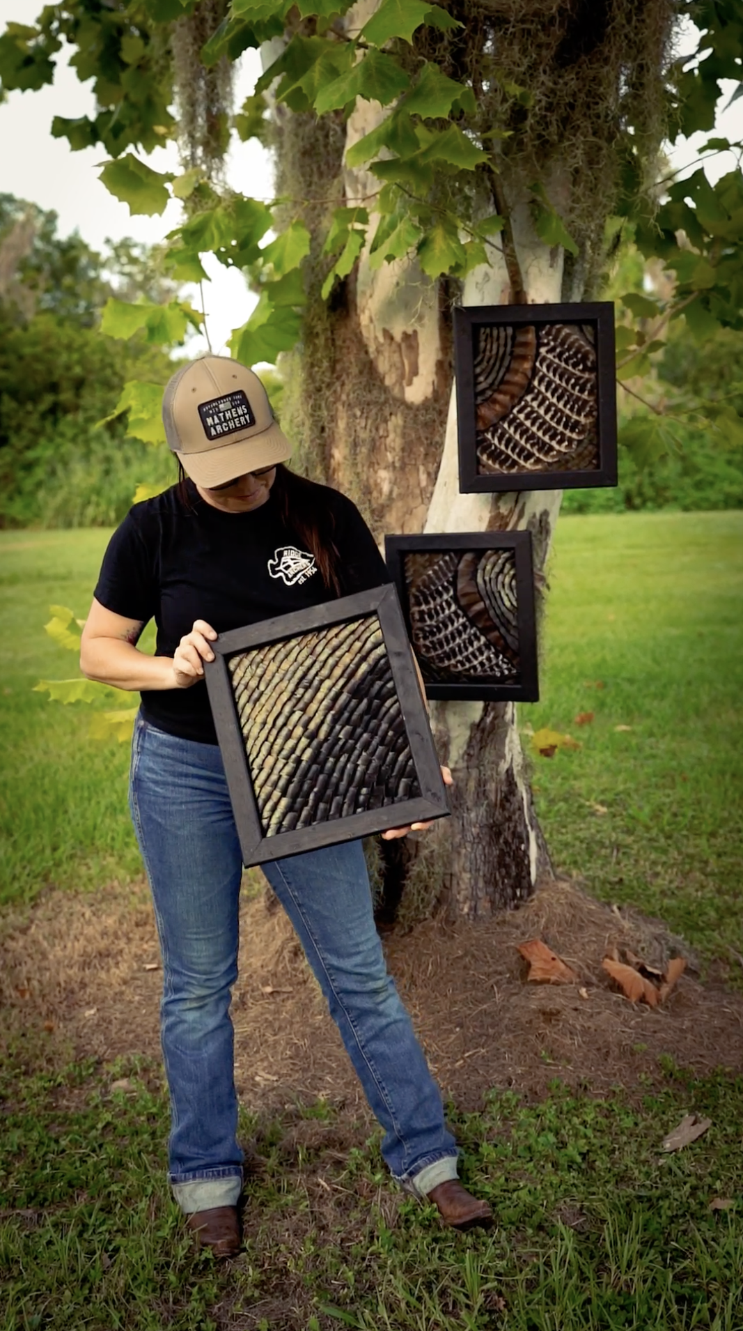 A woman standing outdoors next to a tree displaying three framed art pieces with abstract, circular patterns made of shells or similar natural materials. The woman wears a black T-shirt with a logo, blue jeans, a beige cap, and brown boots.