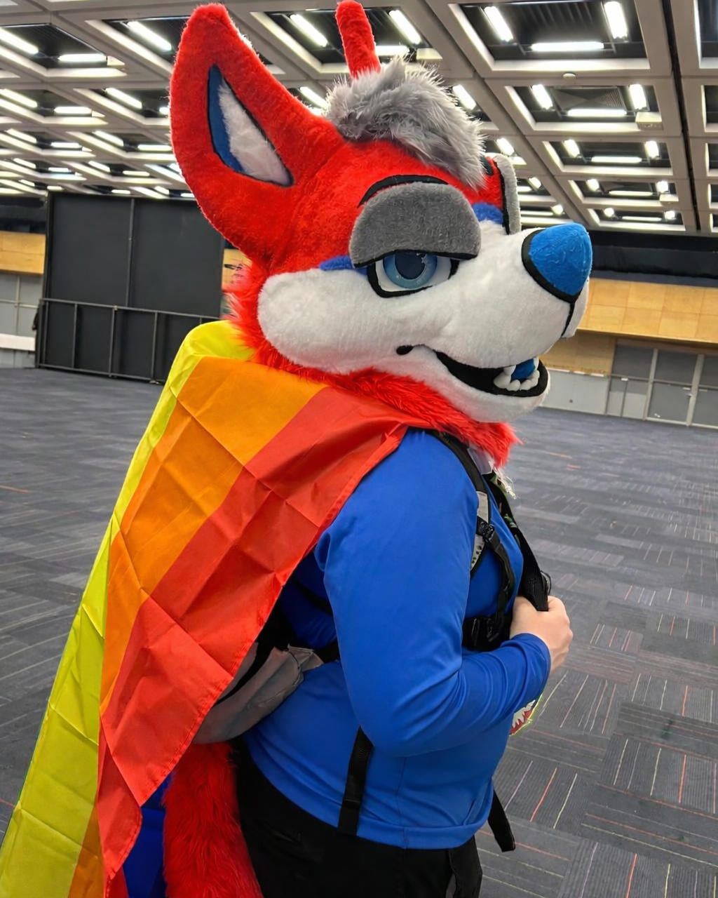 Person in a blue jacket with a furry fox mascot head, large ears, and a colorful cape, standing in an indoor setting with a high ceiling and industrial lighting.
