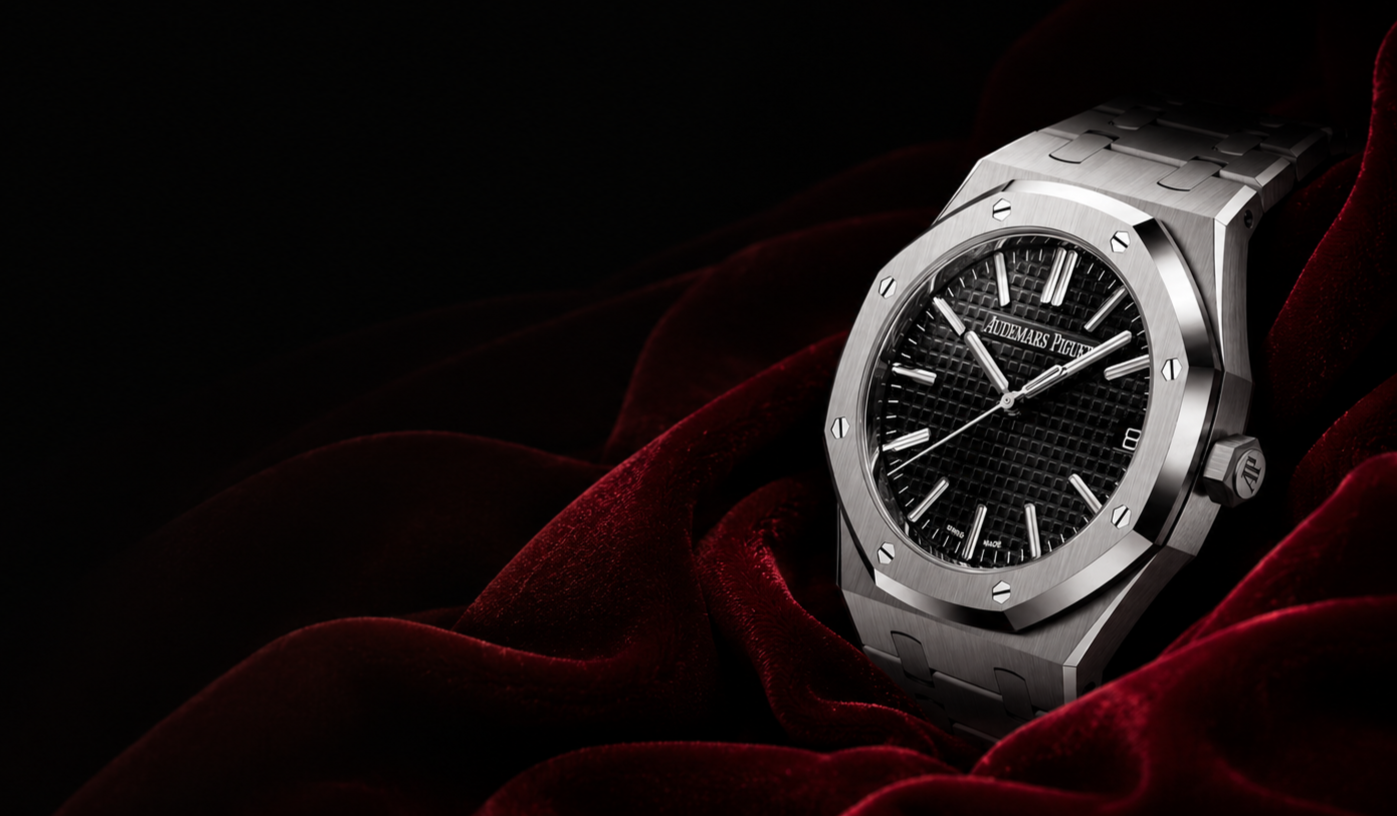 A luxury Audemars Piguet Royal Oak watch with a black textured dial, silver hour markers and hands, set against dark red velvet fabric.