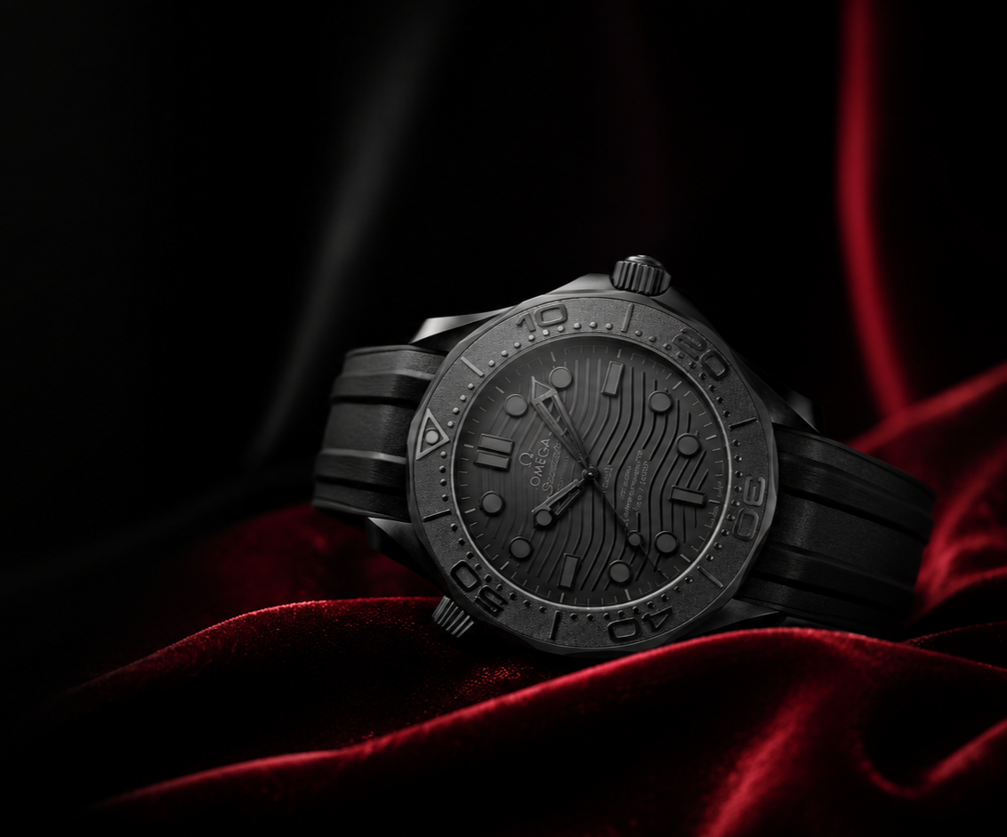 A black Omega Seamaster diving watch resting on red velvet fabric, with a black textured dial and black strap, against a dark background.