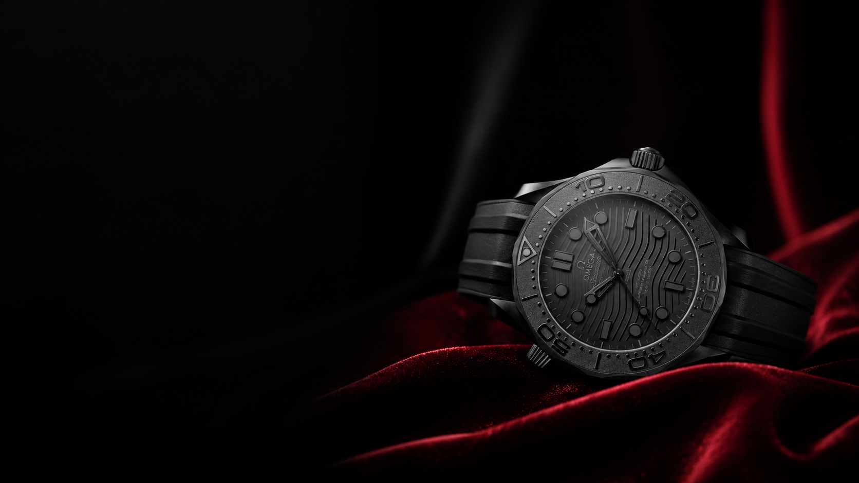 A black Omega Seamaster wristwatch with a textured dial and rubber strap, resting on red velvet fabric against a dark background.