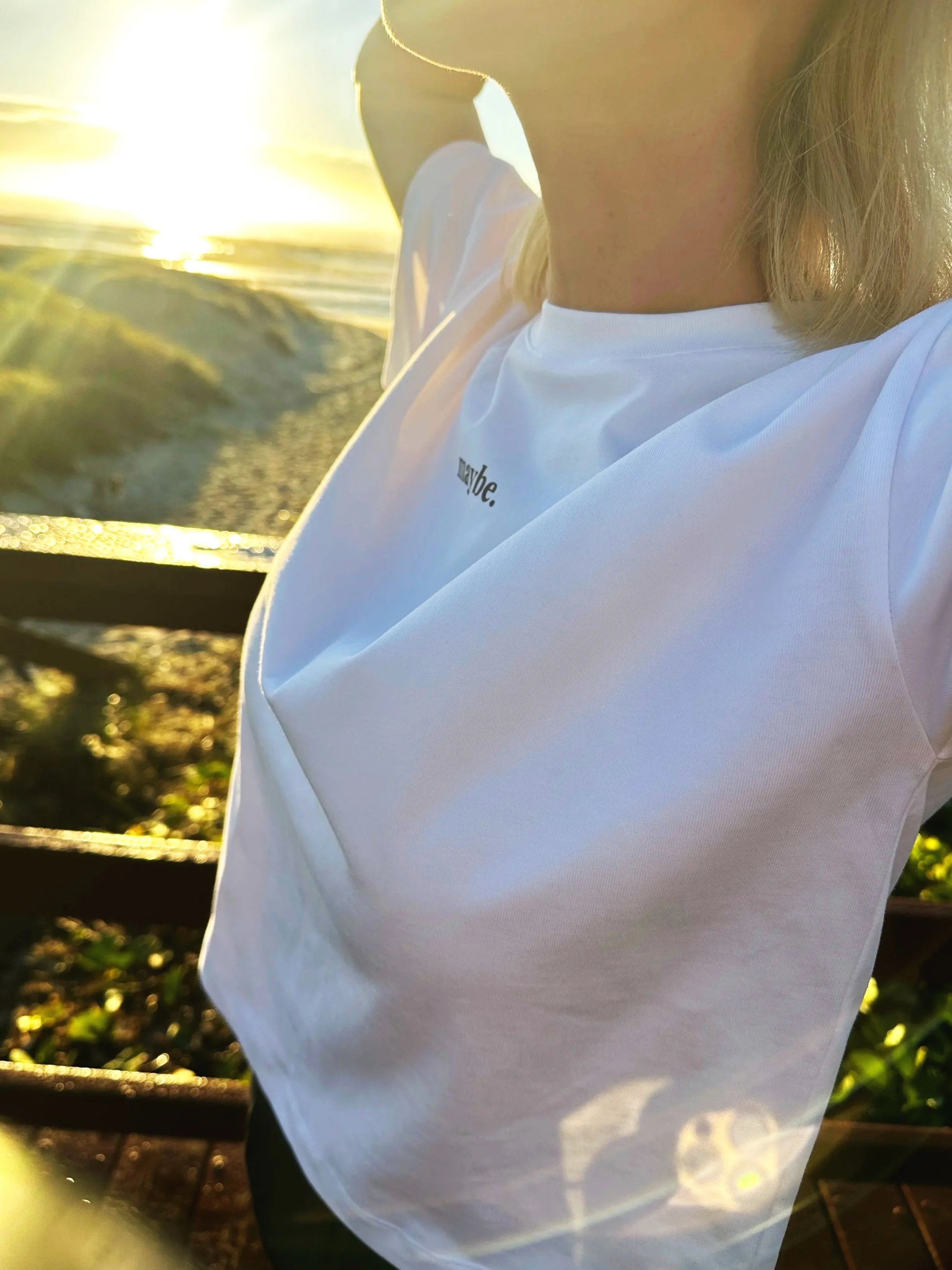 Close-up of a woman with shoulder-length blonde hair wearing a white t-shirt with text, standing outdoors near water with sunlight in the background.
