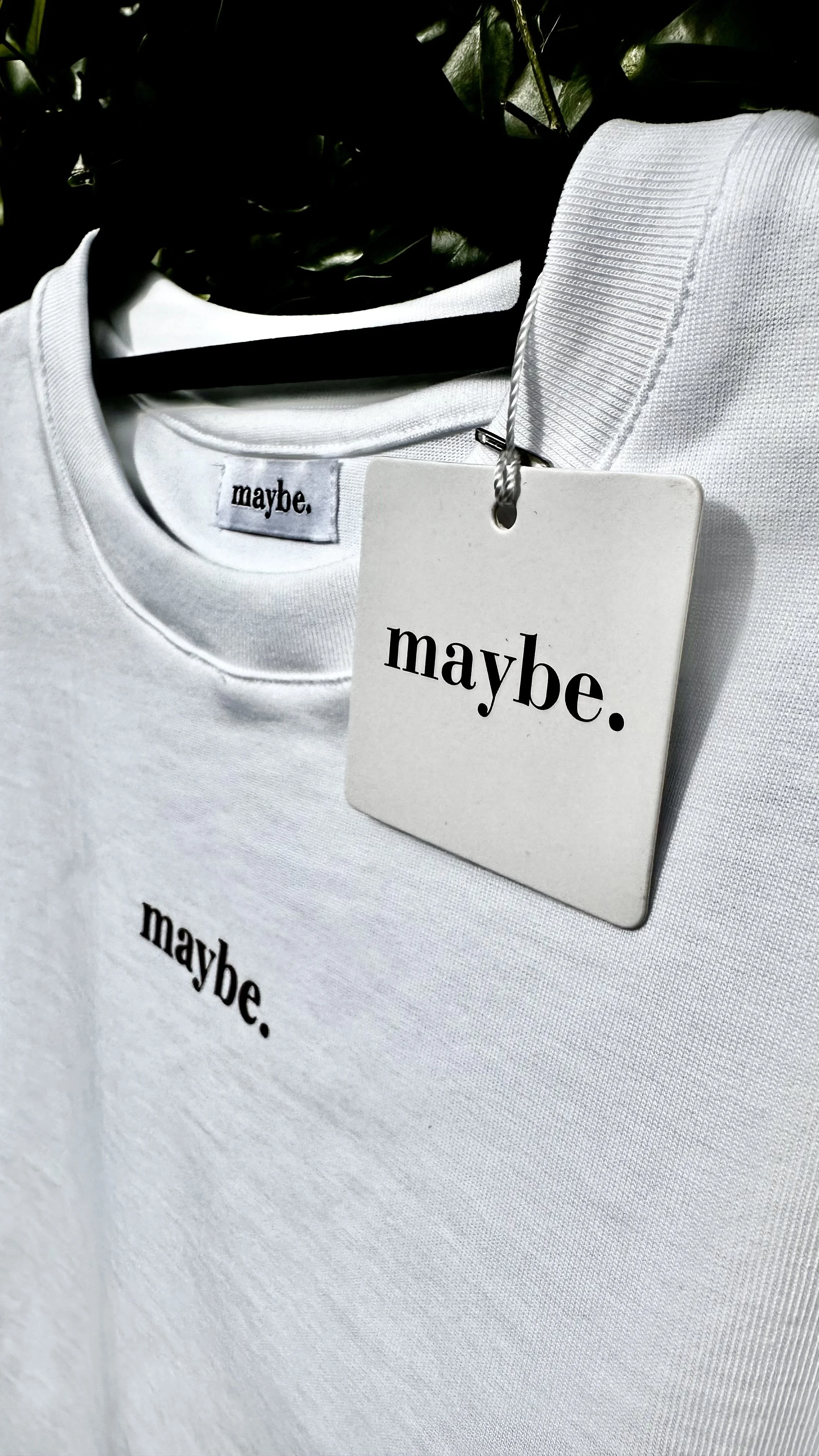 White T-shirt with the word 'maybe.' hanging on a hanger with a clothing tag that also says 'maybe.'