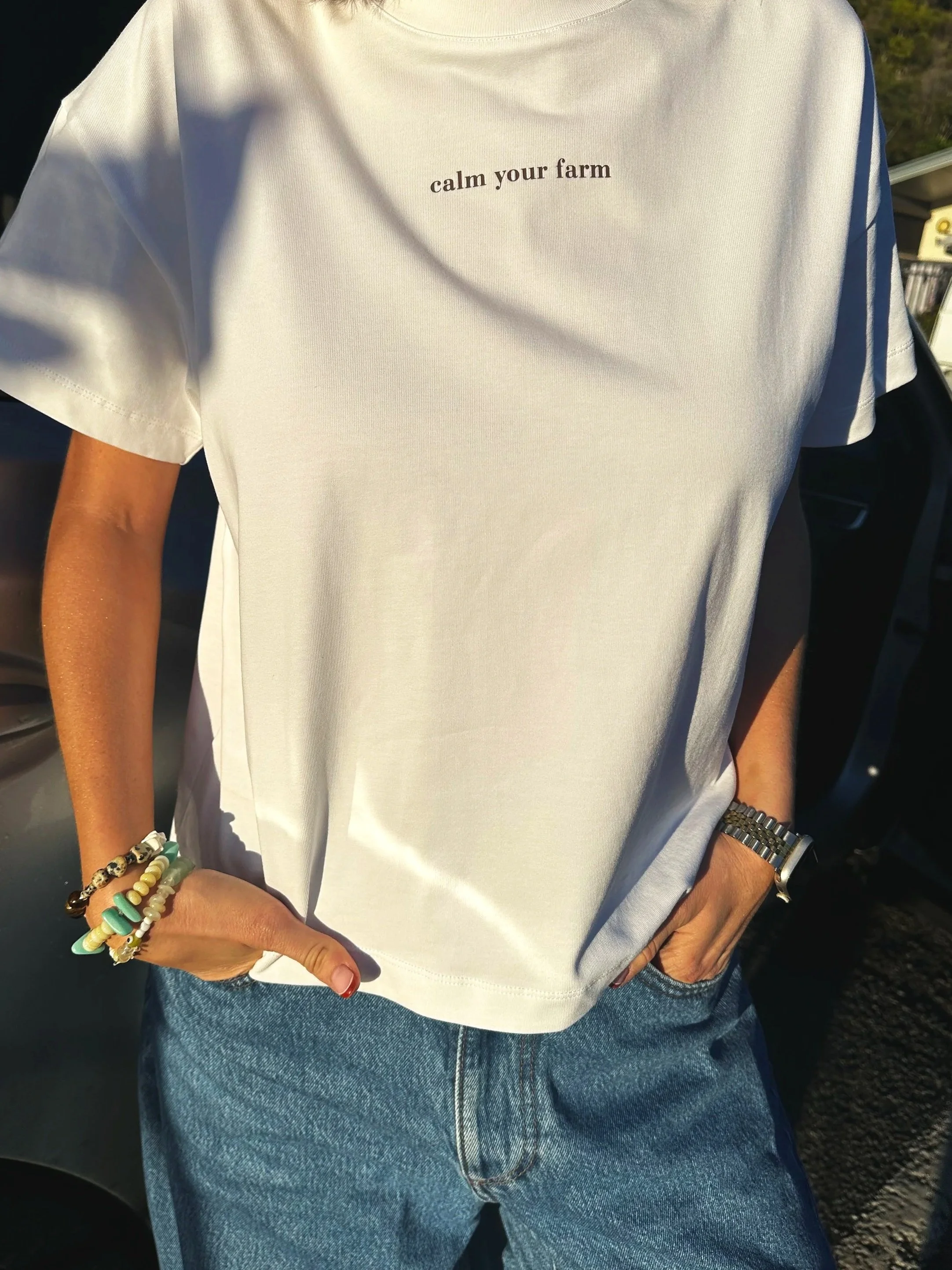 Person wearing a white t-shirt with the text 'calm your farm' on the upper left side, blue jeans, a watch on the left wrist, and beaded bracelets on the right wrist. The person's right hand rests on the right hip. The background shows a car and part of an outdoor setting.