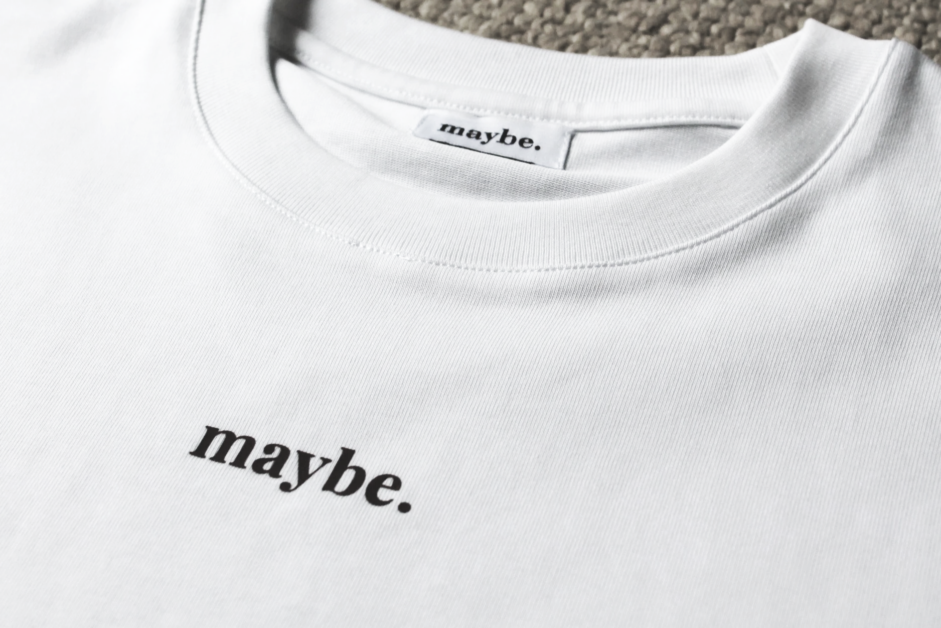 Close-up of a white T-shirt with a partial chocolate text that says 'maybe.' at the neckline, laying on a textured surface.