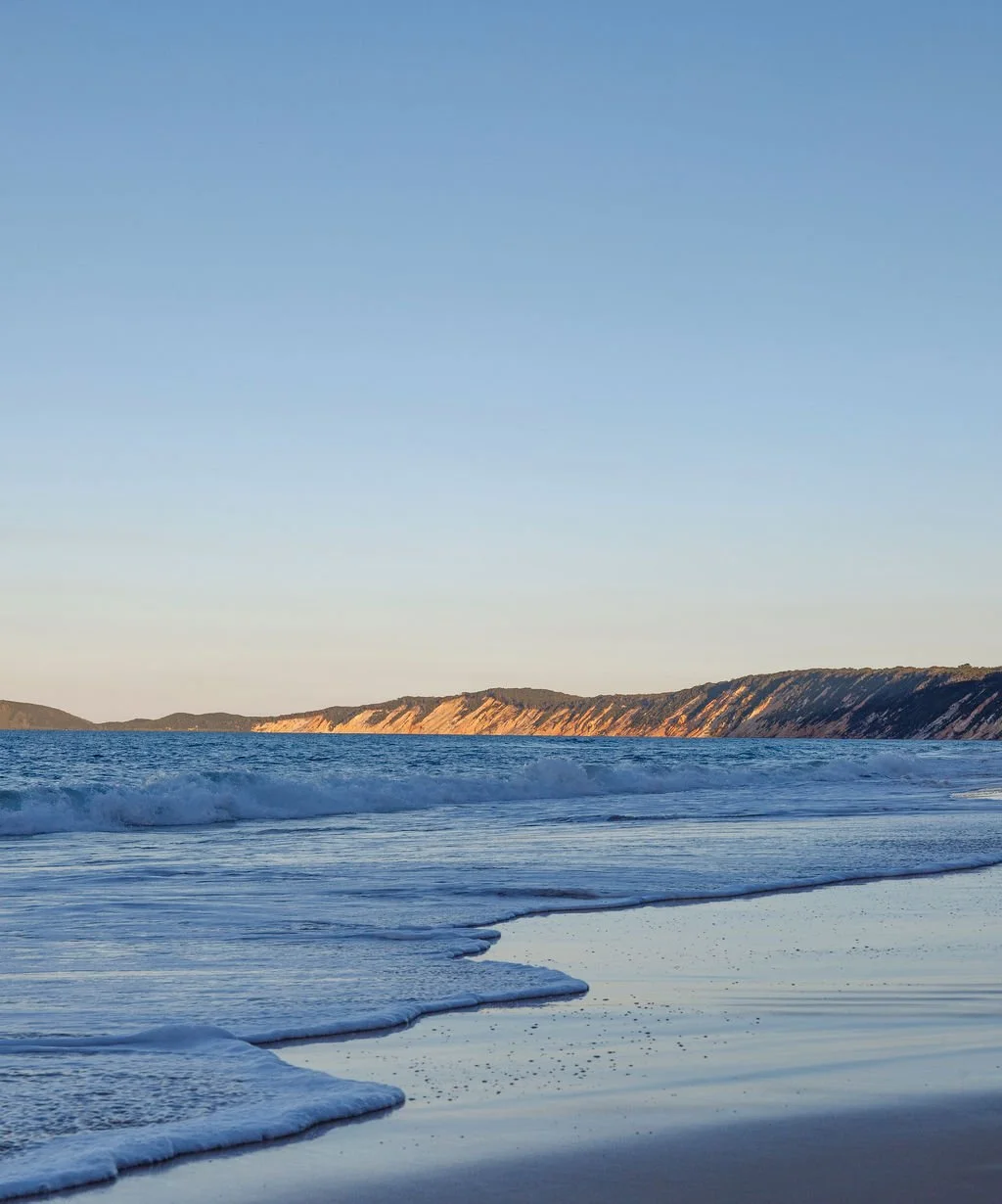 A serene ocean beach scene with gentle waves, golden cliffs in the distance, and a clear blue sky.