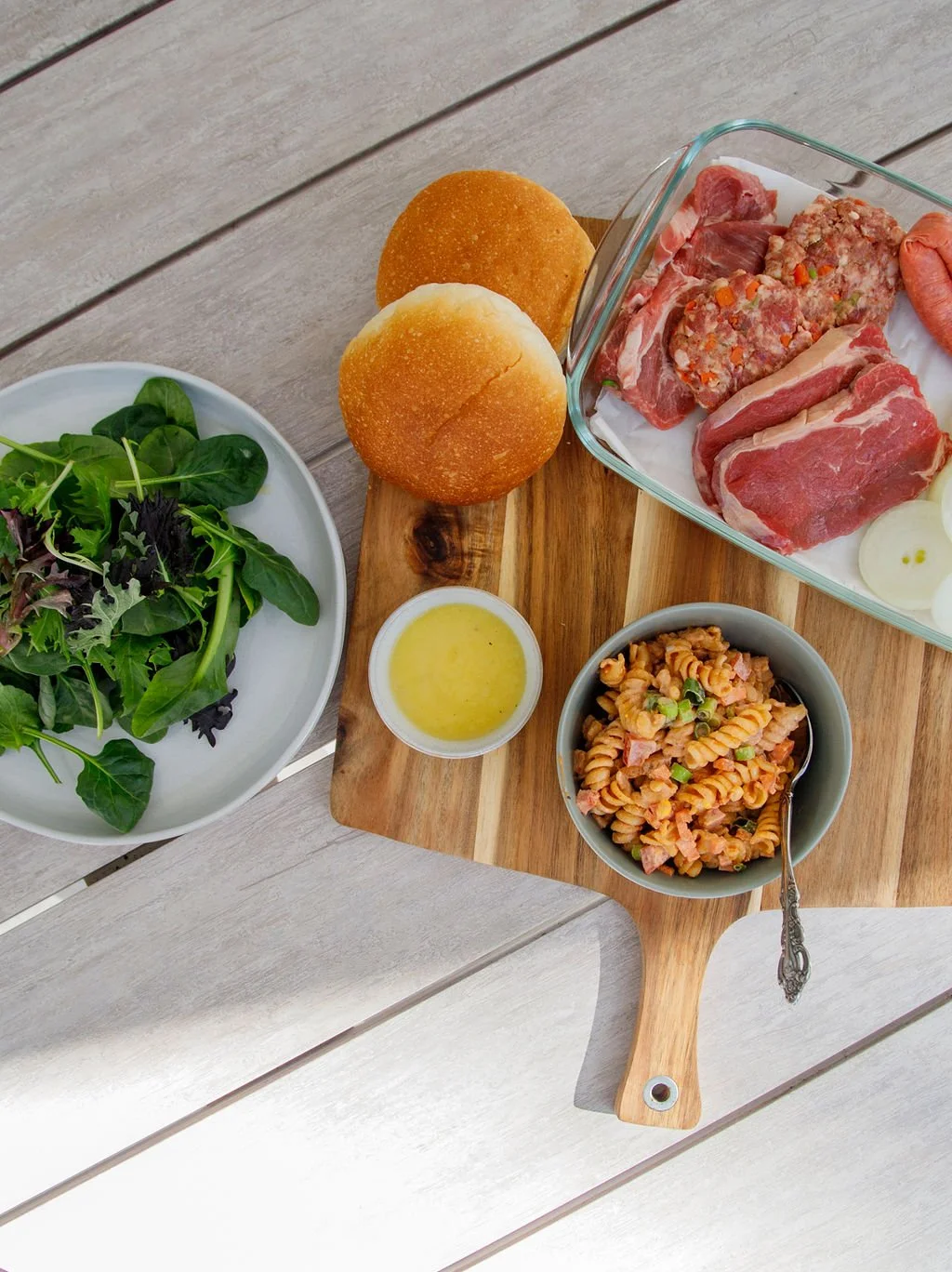 A wooden table holds a bowl of mixed greens, a small dish of olive oil, two bread rolls, a small bowl of pasta salad, and a wooden cutting board with a glass baking dish of raw meat, a sausage, two onion slices, and a small dish of olive oil.