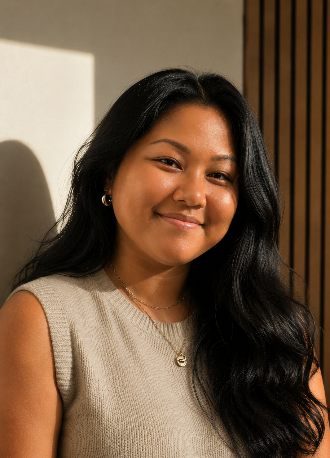 A young woman with long black hair smiling at the camera, wearing a beige sleeveless top and jewelry, standing indoors with a wooden panel background and sunlight casting shadows.