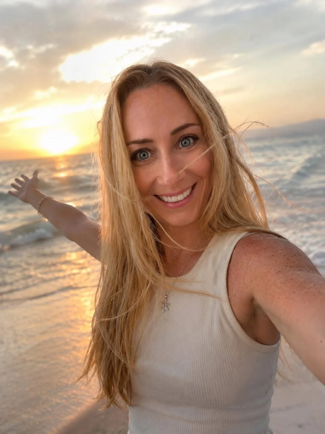 A smiling woman with long blonde hair taking a selfie on the beach at sunset, with her arm outstretched and the ocean and sky in the background.