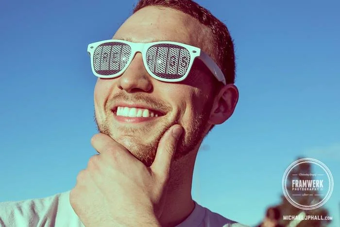 Man wearing white sunglasses with digital numbers, smiling, with hand on chin, outdoors on a sunny day.