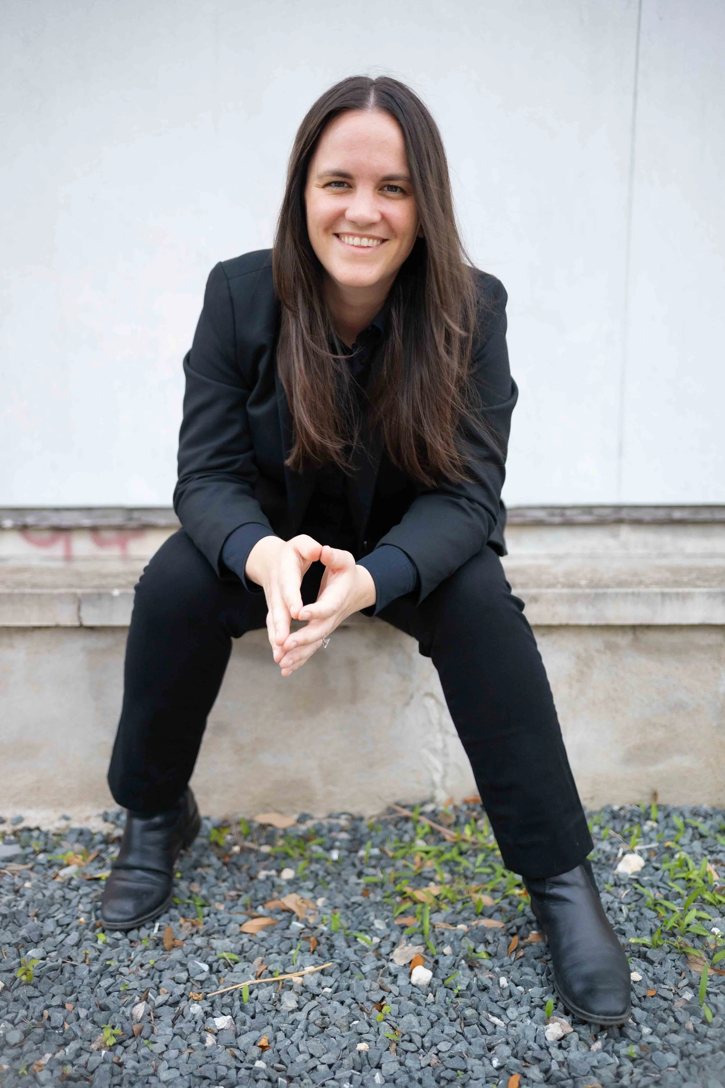 A woman with long dark hair in black clothing sitting on concrete steps, smiling, with a gray wall behind her and small green plants on gravel ground in front.