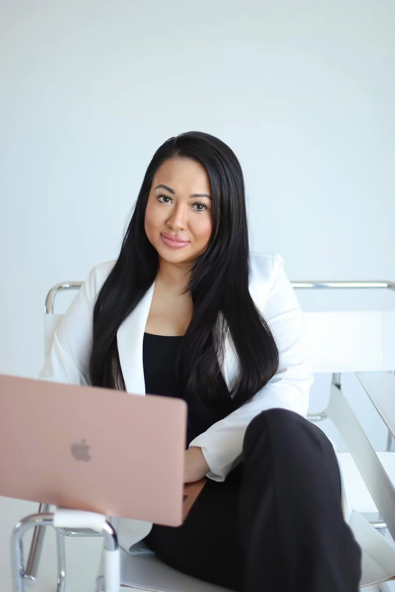 A woman with long black hair, wearing a white blazer, sitting at a desk with a pink laptop in front of her, in a bright, modern office with a blue-gray wall.