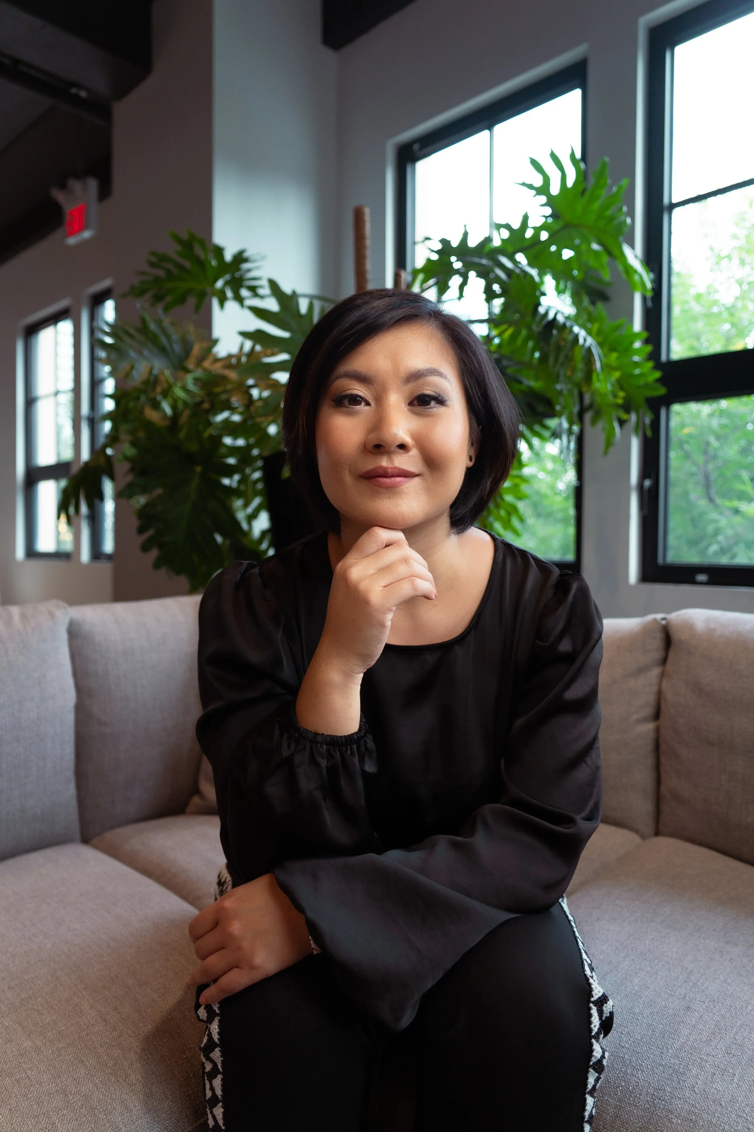 A woman with short black hair and a black top sitting on a beige couch in a modern room with large windows and green indoor plants.