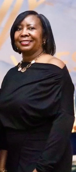 A woman with shoulder-length black hair, wearing a black off-shoulder top and gold jewelry, smiling at the camera.