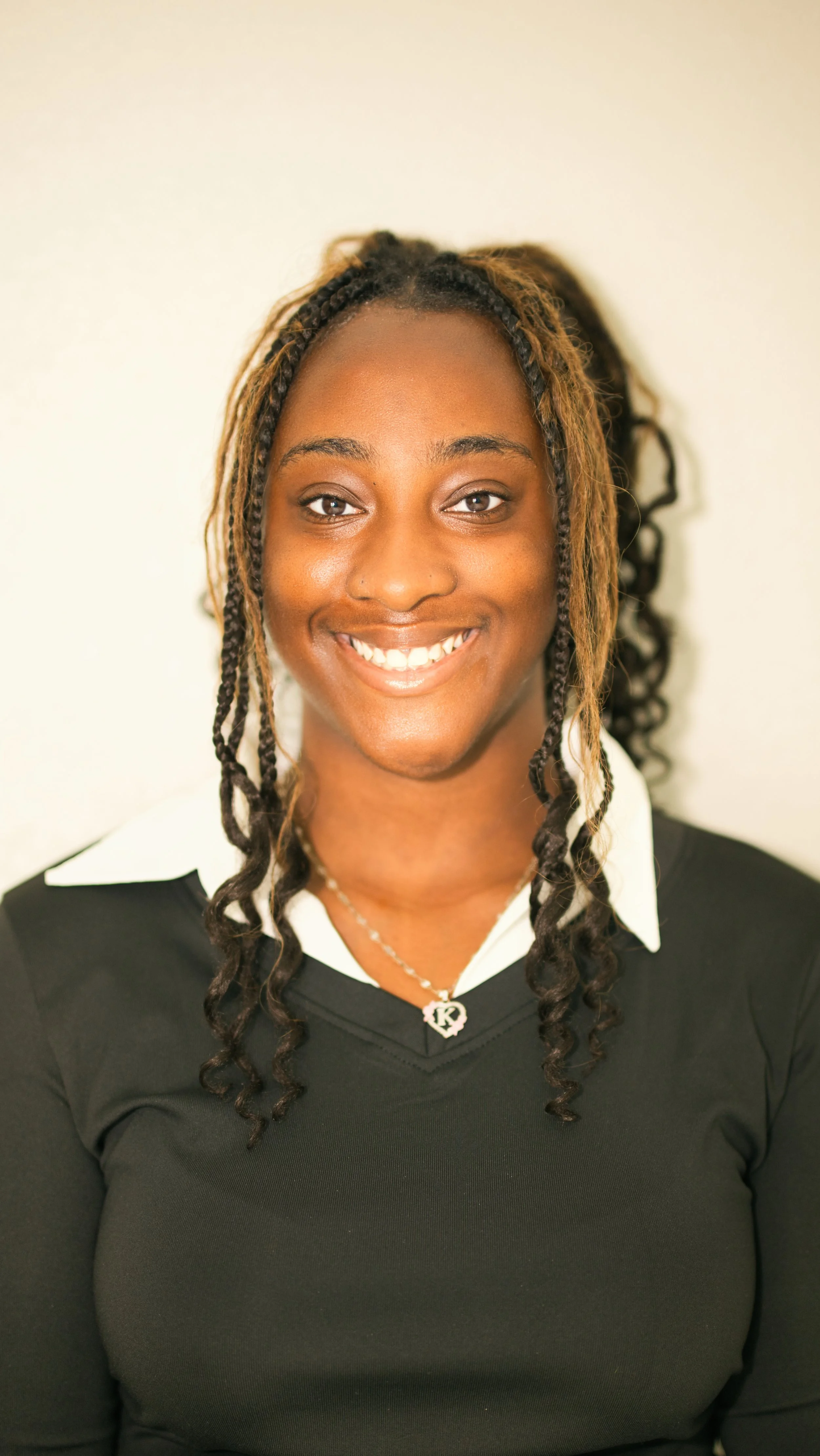 A young woman with long, curly hair styled in braids and curls, smiling at the camera, wearing a black sweater over a white collared shirt, and a silver necklace with a pendant.