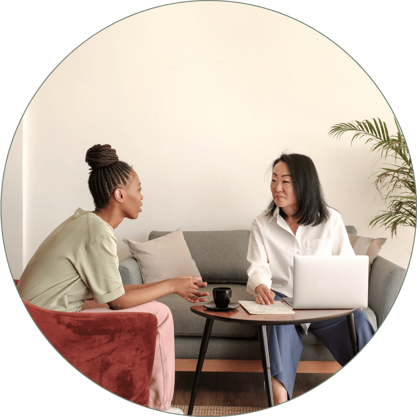 Two women having a discussion in a cozy living room, one sitting on a gray couch with a laptop, the other on a red armchair, with a small round coffee table between them holding a black mug and a notebook.