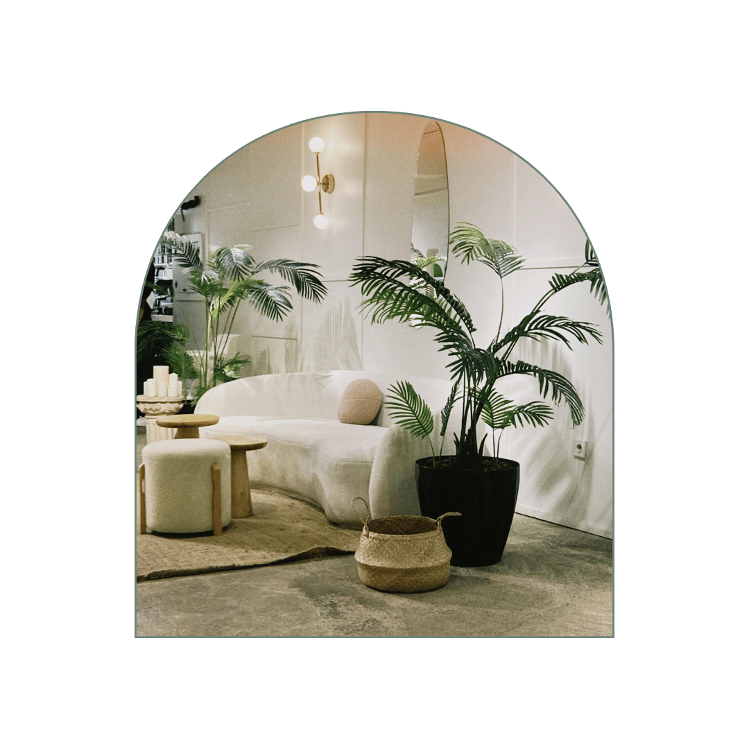 Interior of a modern, minimalistic living room with a white curved sofa, large potted plants, a small beige ottoman, a woven basket, and ambient lighting fixtures.