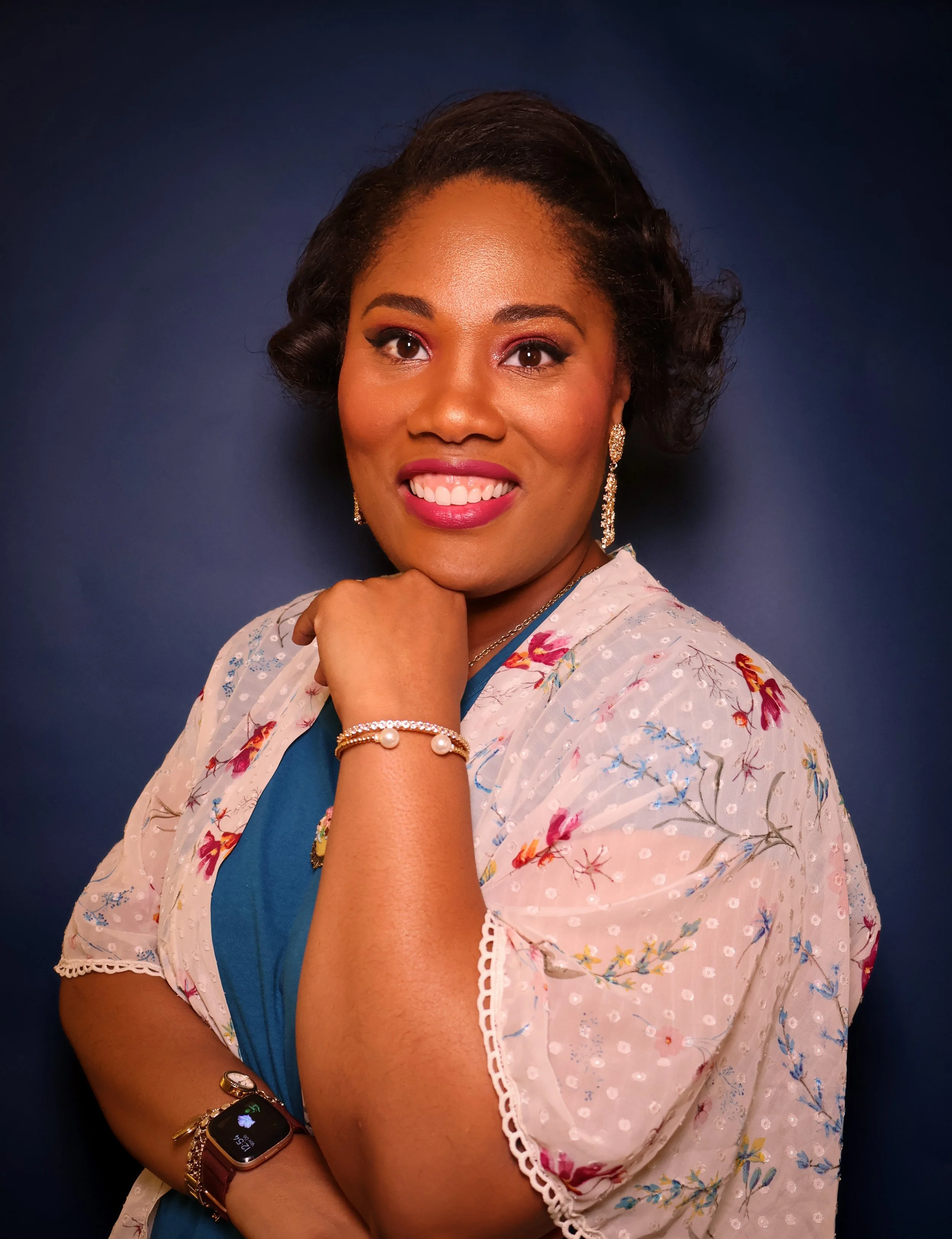 A woman with dark brown skin and short, curled hair is smiling and looking directly at the camera. She is wearing makeup, earrings, a bracelet, and a smartwatch. She is dressed in a floral-patterned blouse with white lace trim at the sleeves and a blue sari with a floral border. Her right arm is crossed over her chest, and her left hand rests near her chin, with her fingers touching her face against a dark blue background.