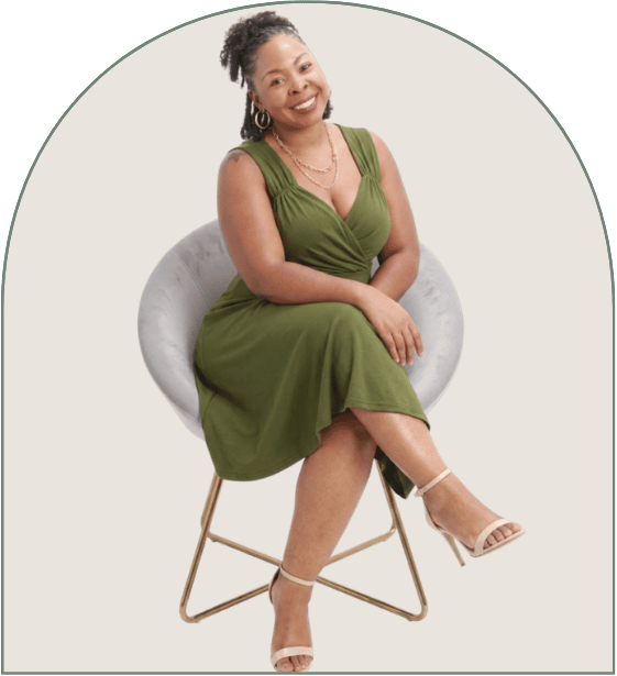 A woman sitting on a modern gray chair with wooden legs, wearing a green dress and white high-heeled sandals, smiling with her arms resting on her lap against a plain white background.