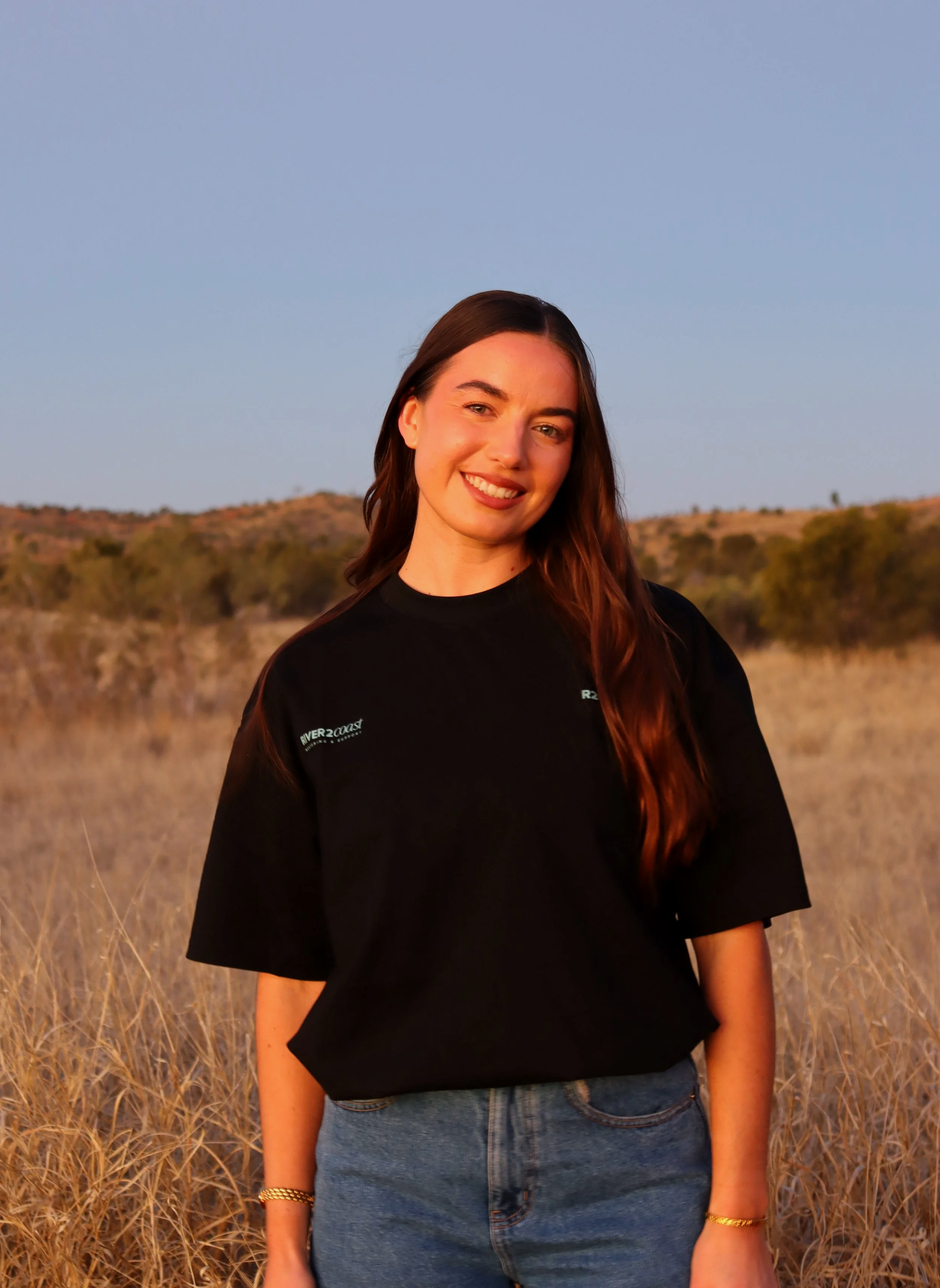A young woman with long brown hair smiling in a field of dry grass during golden hour, wearing a black shirt with text on the sleeve and blue jeans.