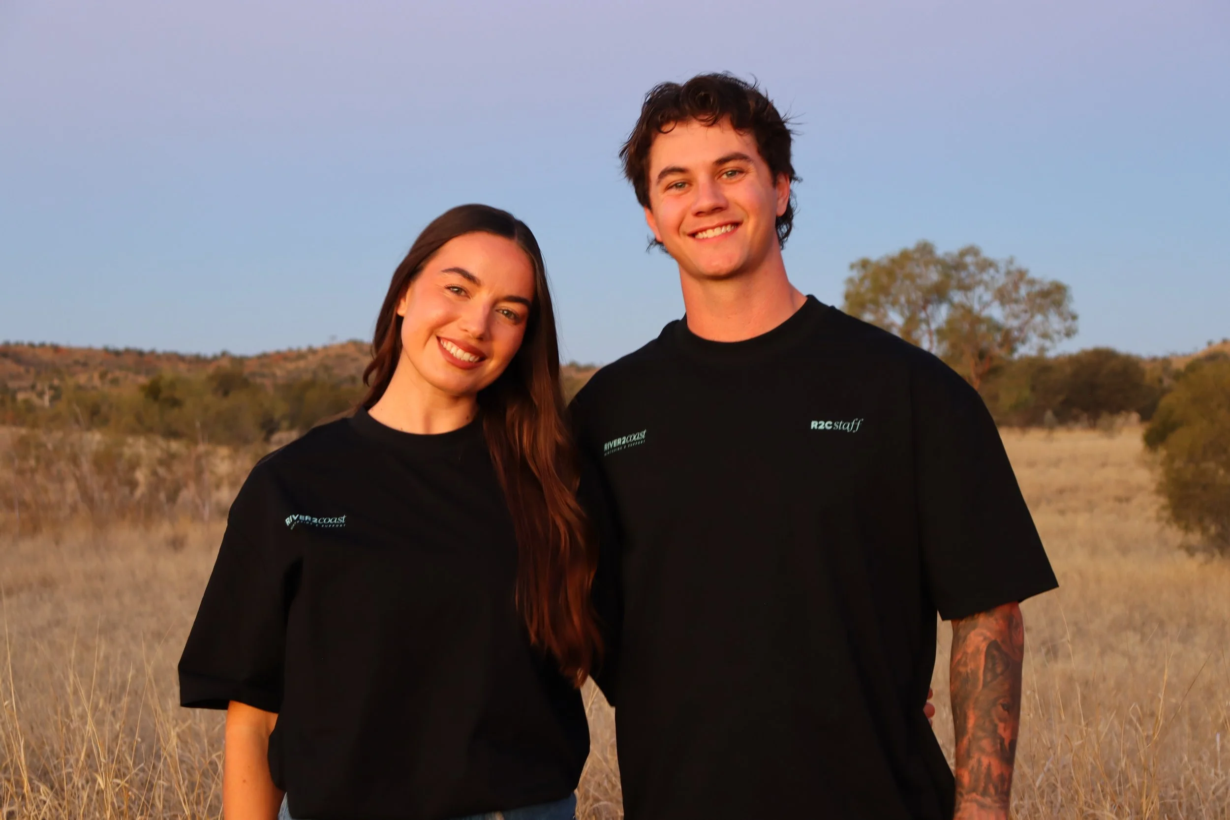 A young woman and a young man smiling outdoors in a field at sunset, wearing black shirts with white text.