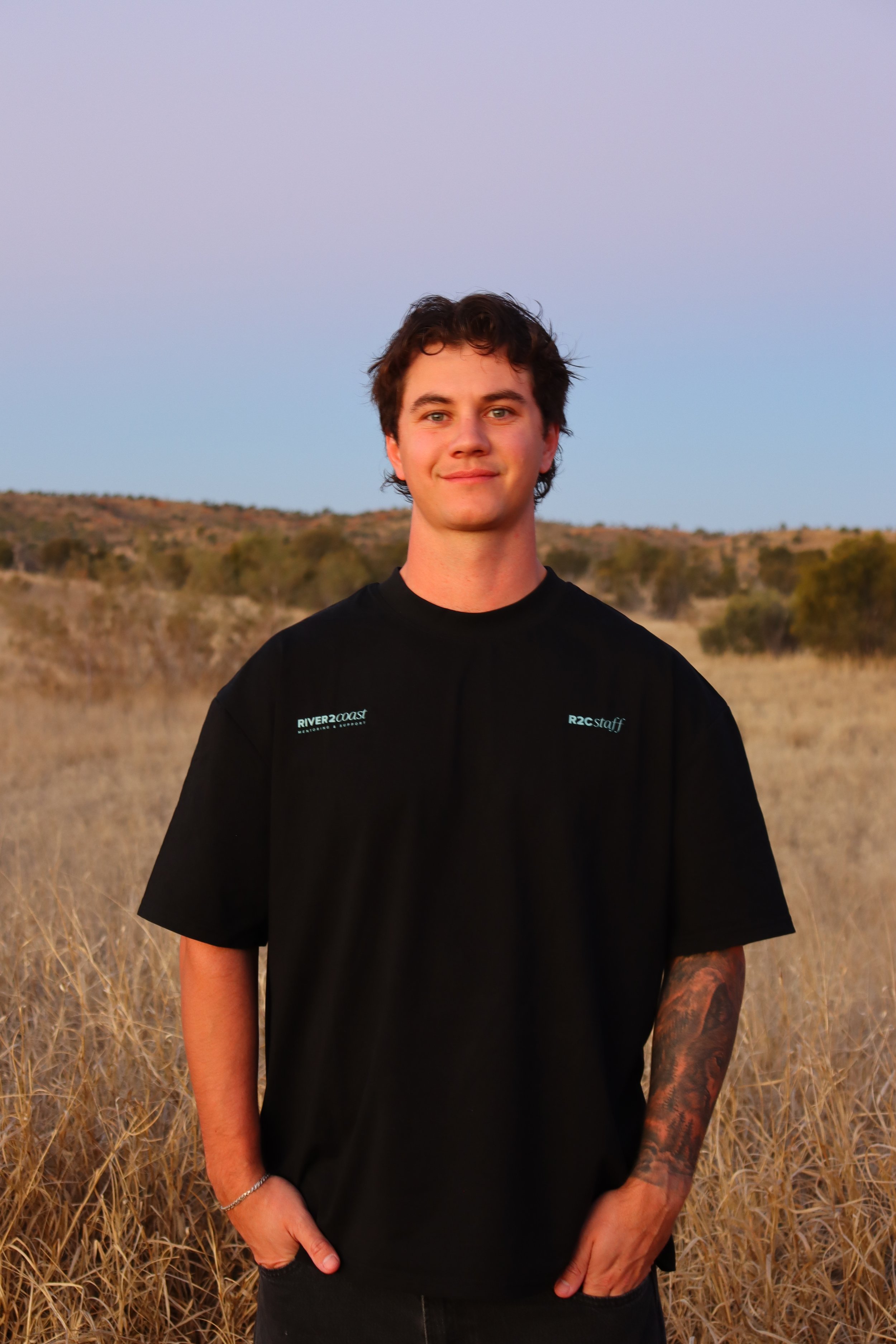 A young man with dark wavy hair and tattoos on his left arm standing in a field of tall, dry grass with hills and trees in the background, during sunset or sunrise.