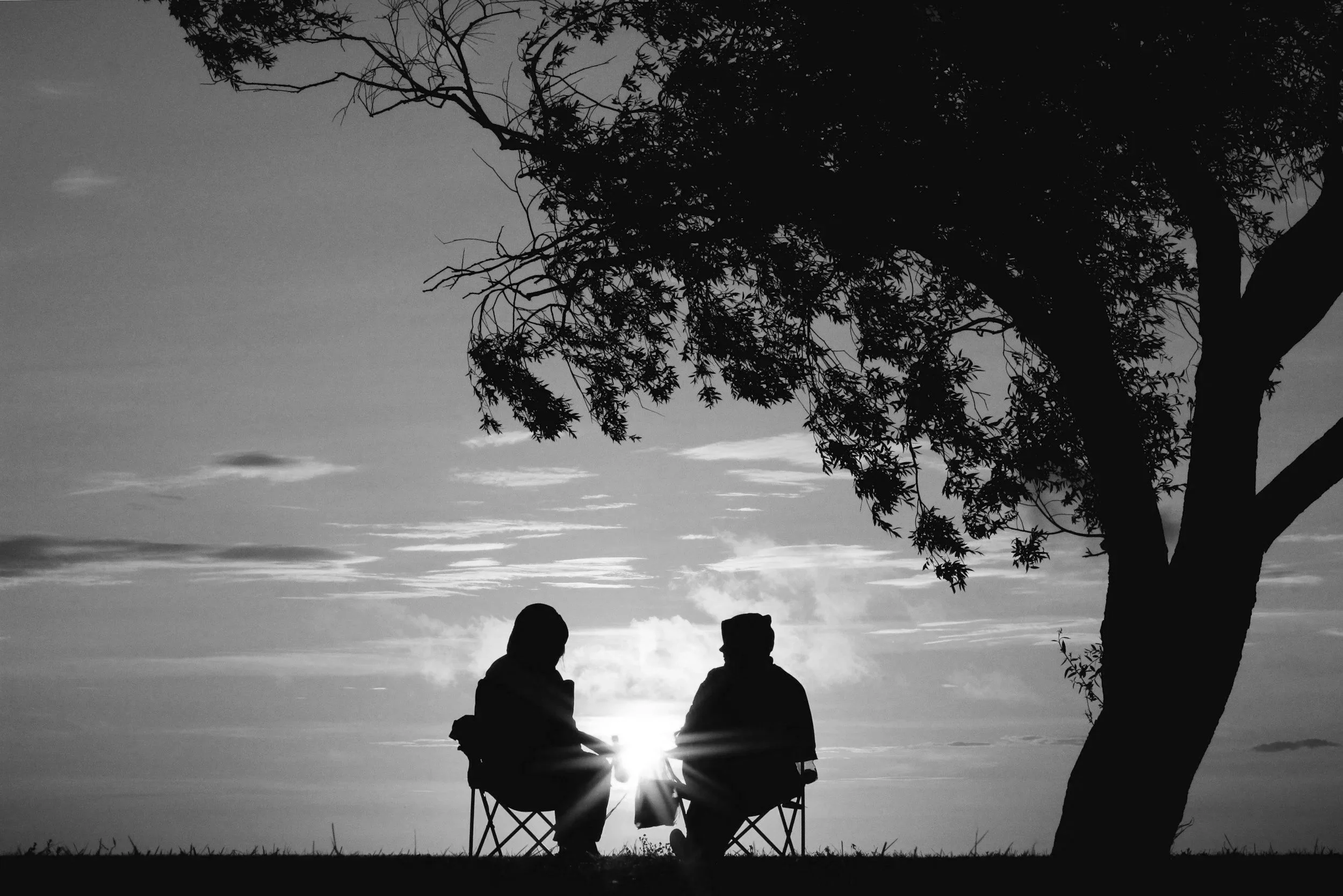 Silhouette of two people sitting in a folding chair outdoors during sunset, with a large tree on the right side and clouds in the sky.