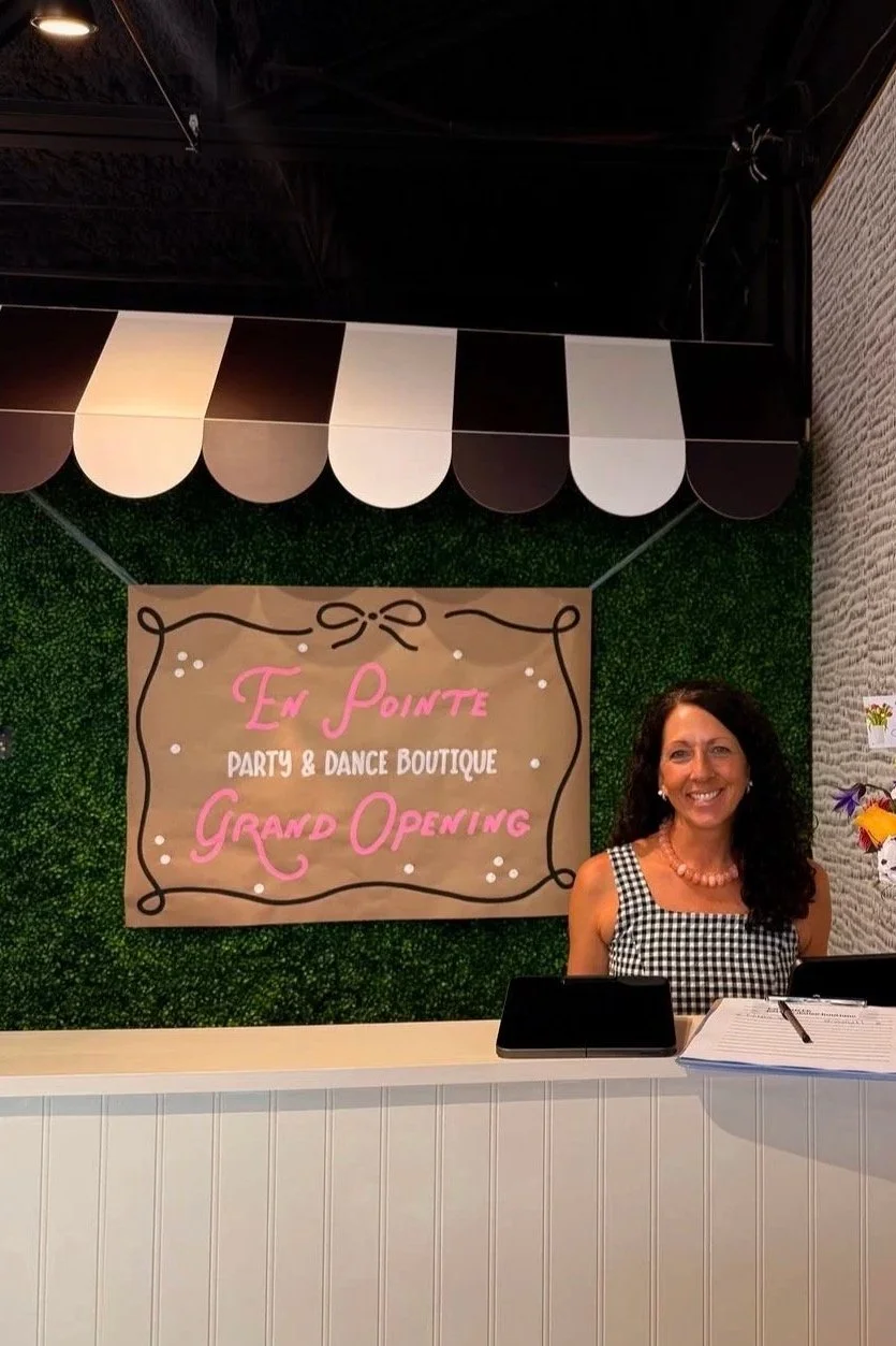 A woman with dark curly hair, wearing a sleeveless black and white checkered dress and a pink necklace, standing behind a reception desk at a boutique grand opening event. Behind her is a sign that reads 'En Pointe Party & Dance Boutique Grand Opening' and a decorative wall with green foliage and a black and white striped canopy.