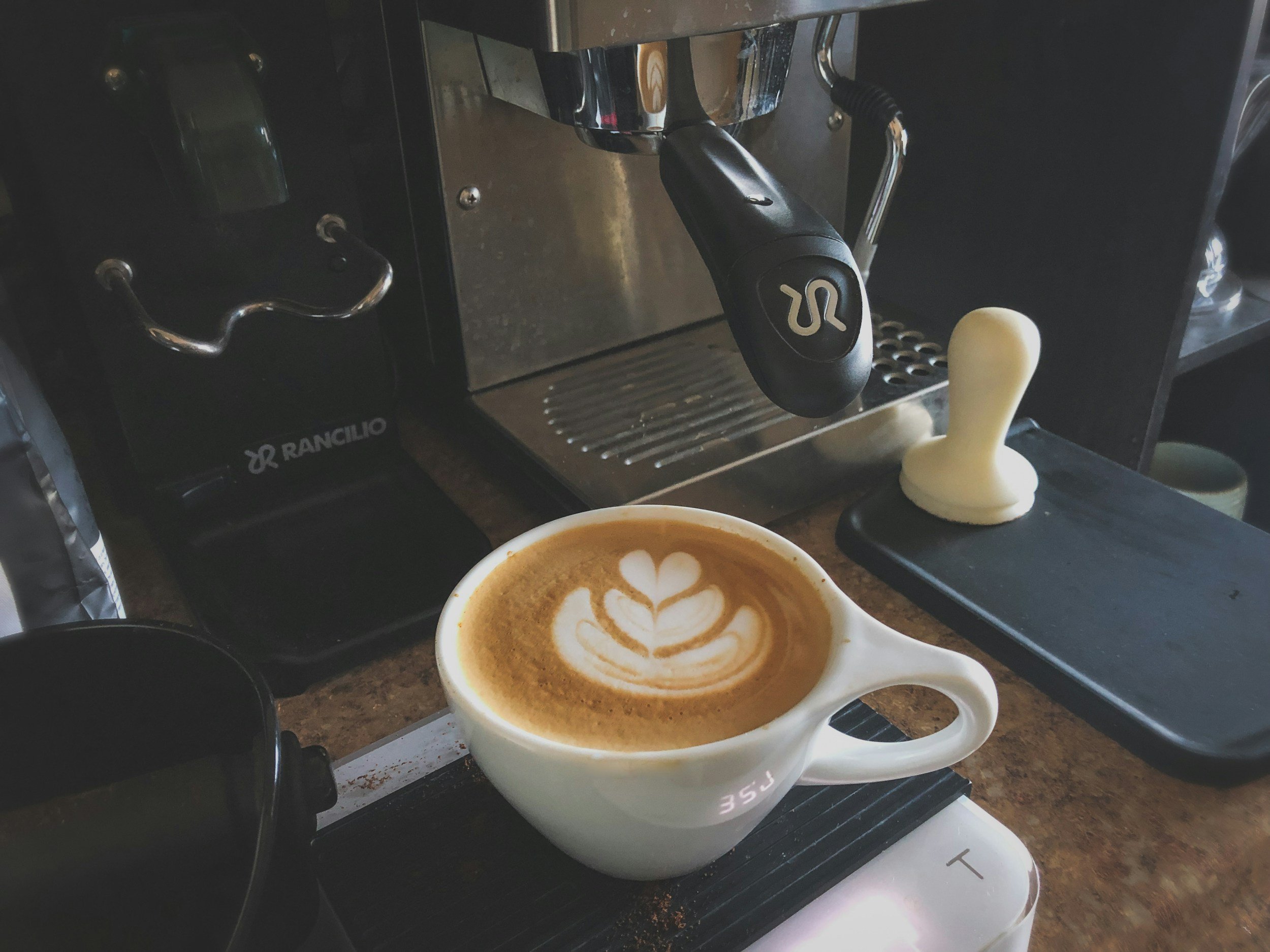 A coffee machine with a portafilter attached, brewing a cappuccino with latte art in a white mug below, on a kitchen countertop.