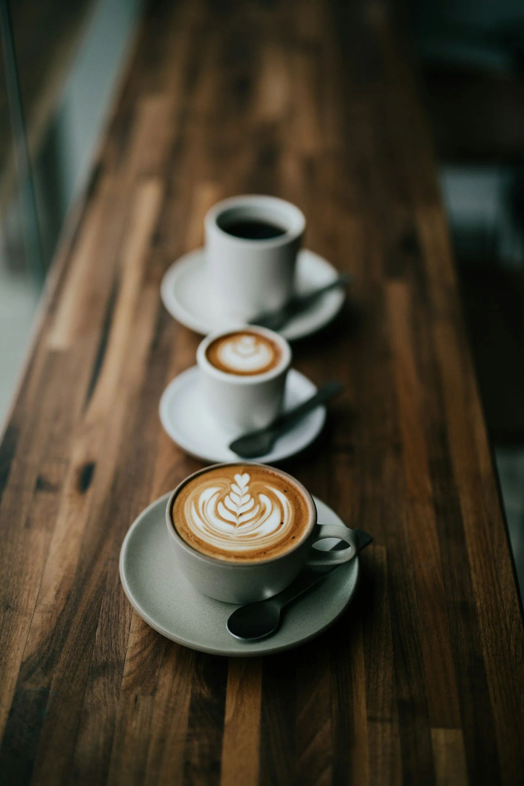 Three cups of coffee with latte art on a wooden table, with spoons on saucers in a cafe setting.