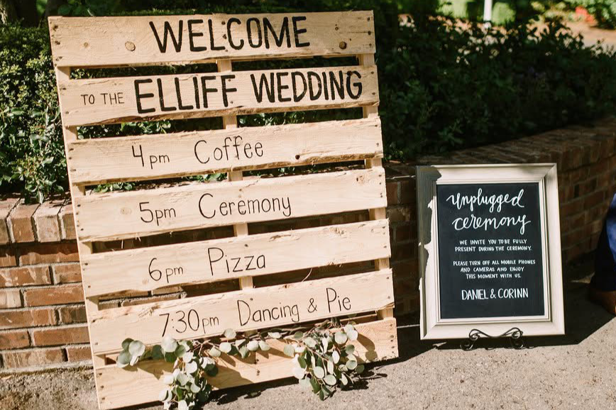 Wooden wedding schedule board  painted on a wood pallet board with events and times listed, next to a framed sign for unplugged ceremony, outdoors with greenery and brick wall.