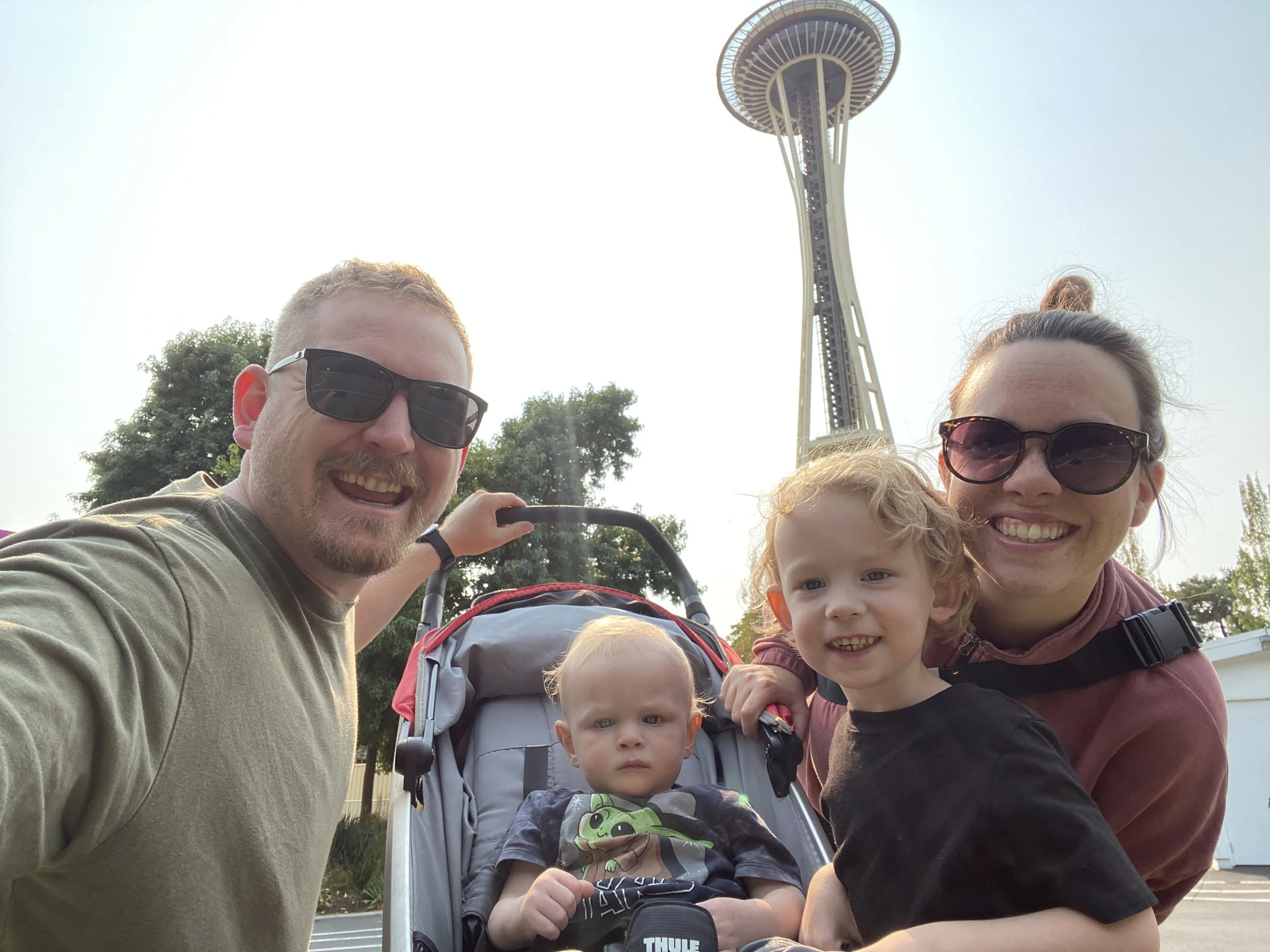 A family of four smiling outdoors in front of the Seattle Space Needle, including a woman, two young children, and a man taking their selfie.