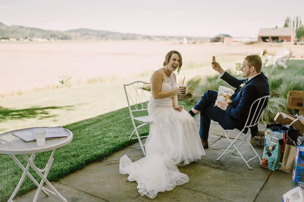 A bride and groom sitting outdoors, with the bride laughing and holding a drink, and the groom giving a toast with a small dessert in hand. There are chairs, a small table with a drink, and various gift bags nearby, with a rural landscape in the background.