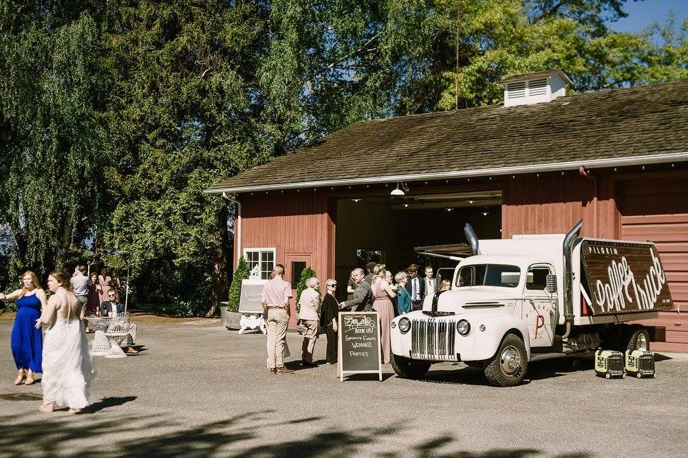 A vintage truck serving as a mobile cafe, parked outside a red barn during a wedding gathering surrounded by trees. Several people are mingling and walking around, with some sitting on benches, at an outdoor wedding on a sunny day.