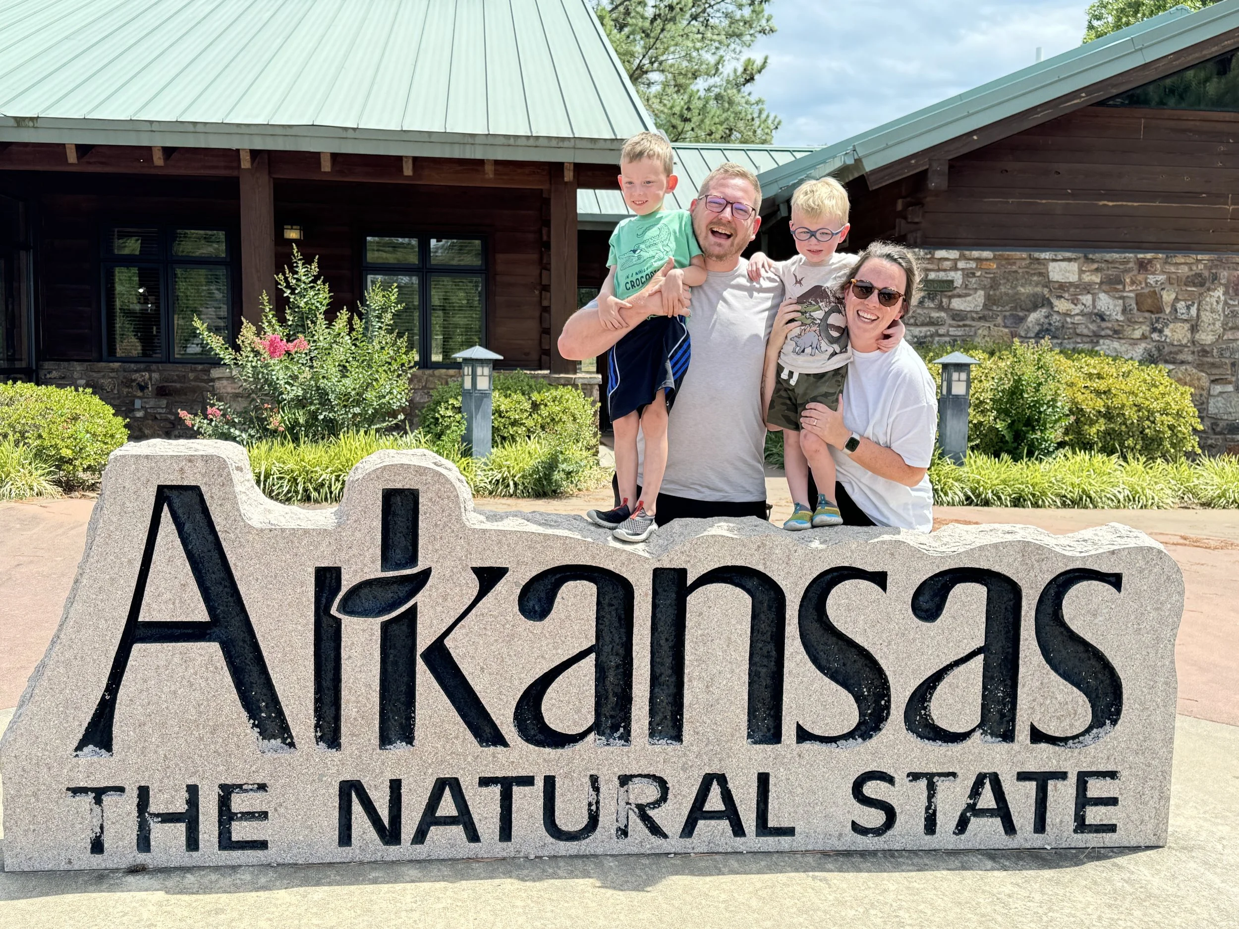 A family of four standing together outdoors in front of a building with a green metal roof and stone wall, with greenery and trees behind. They are smiling and hugging, with two young boys and their parents. A large stone sign in the foreground reads "Arkansas the Natural State."